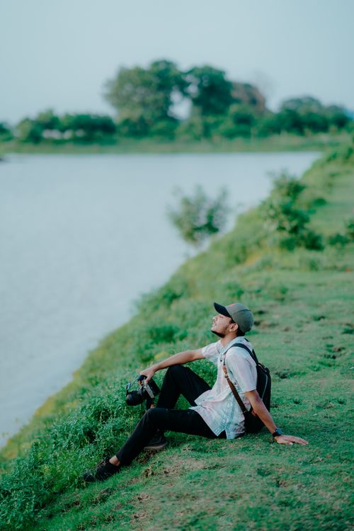 Free A man sits thoughtfully on a grassy riverbank, enjoying a peaceful day outdoors. Stock Photo