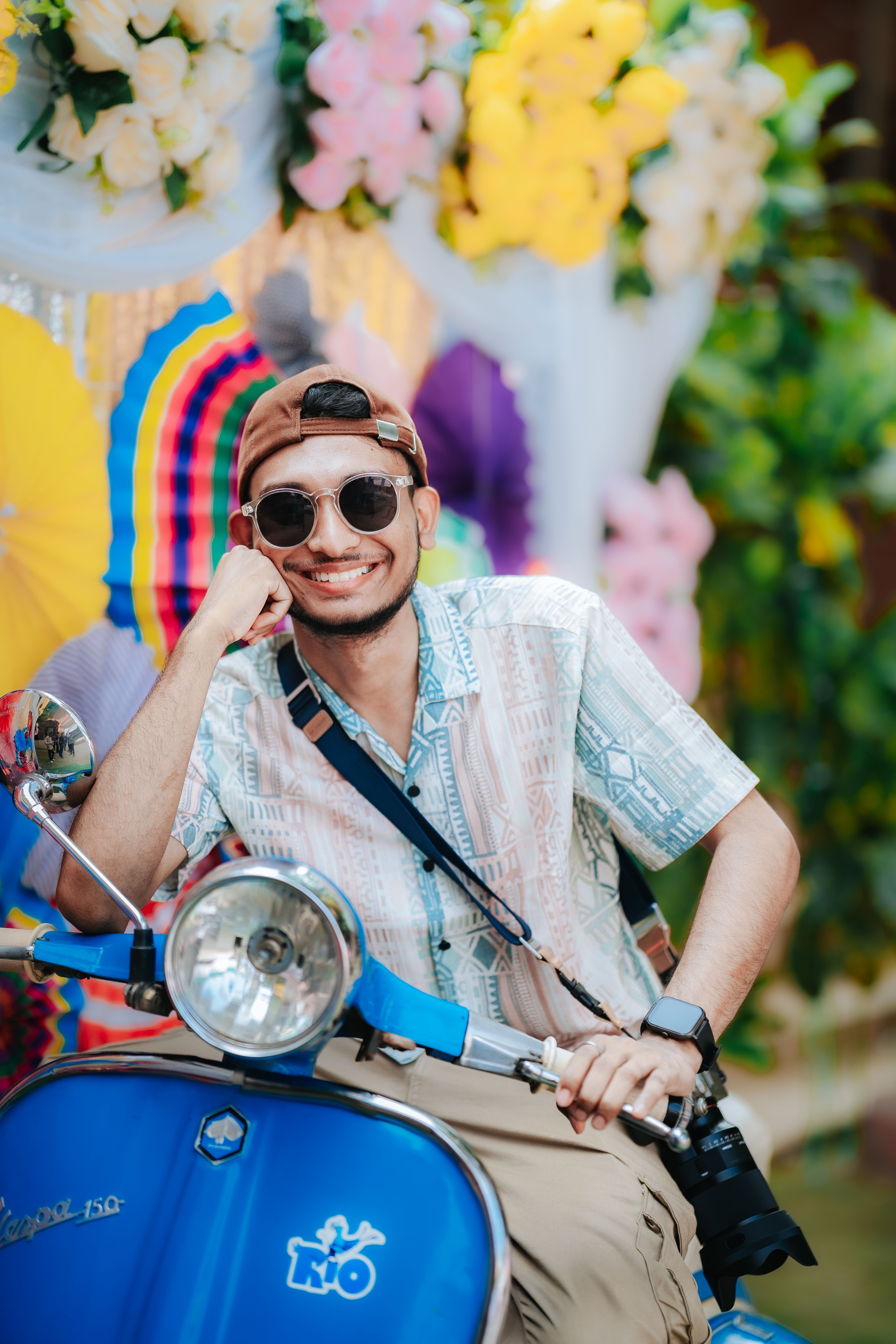Smiling man in sunglasses posing on scooter with colorful background at a summer festival.
