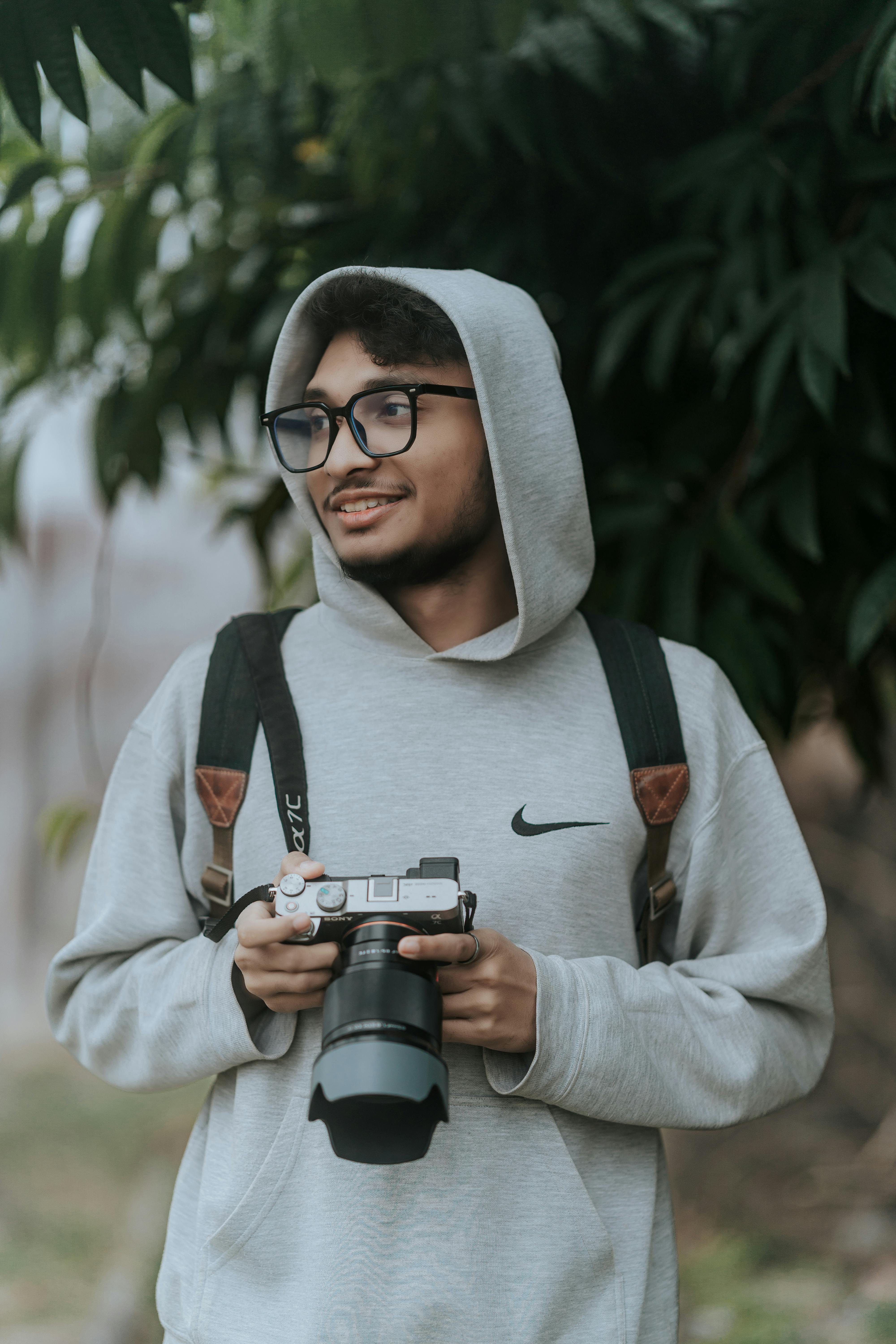 Kostenlos Ein junger Mann mit Brille hält im Freien eine Kamera und trägt einen Kapuzenpullover. Stock-Foto