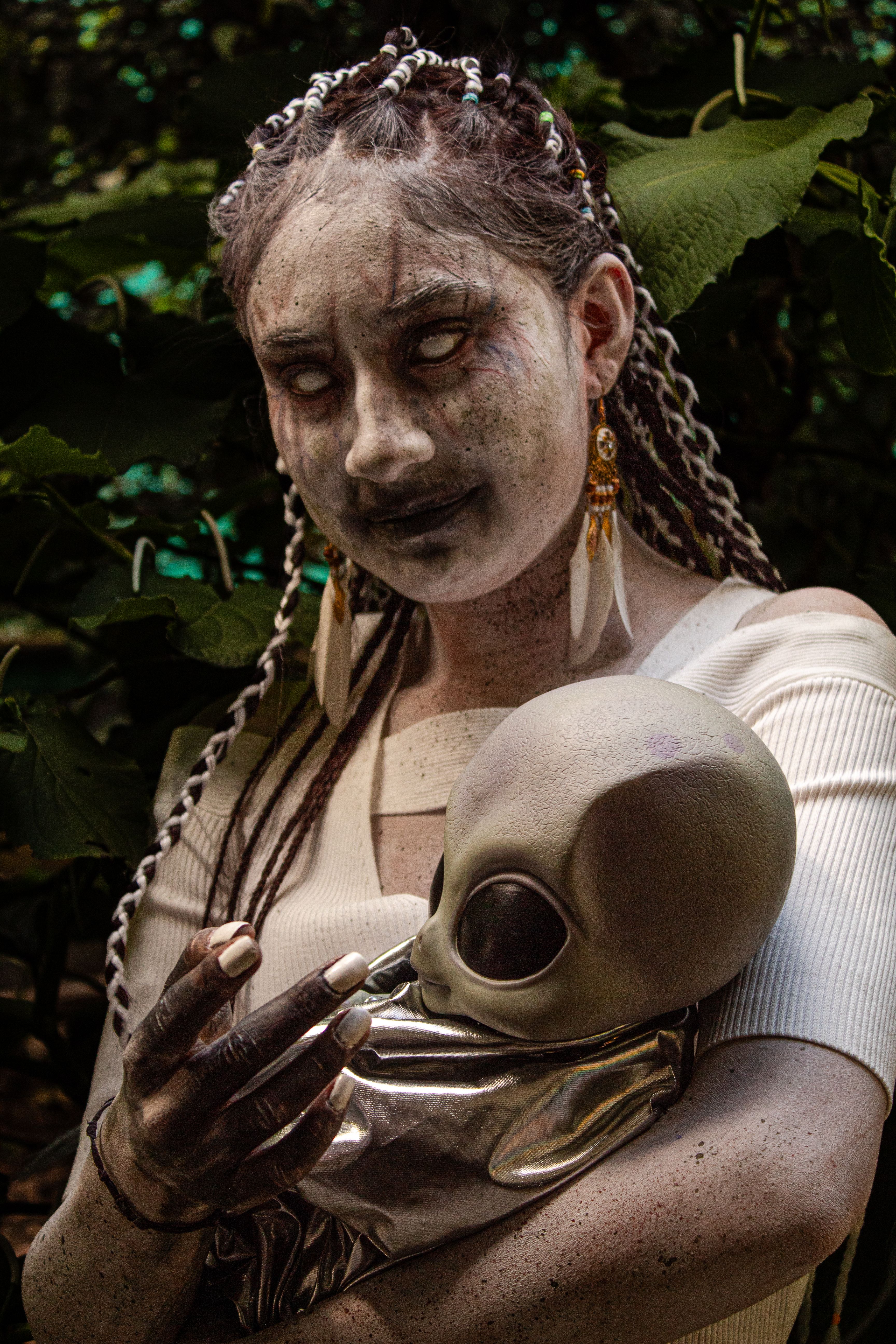 A woman in gothic attire with braided hair and face paint holds a mysterious alien doll outdoors.