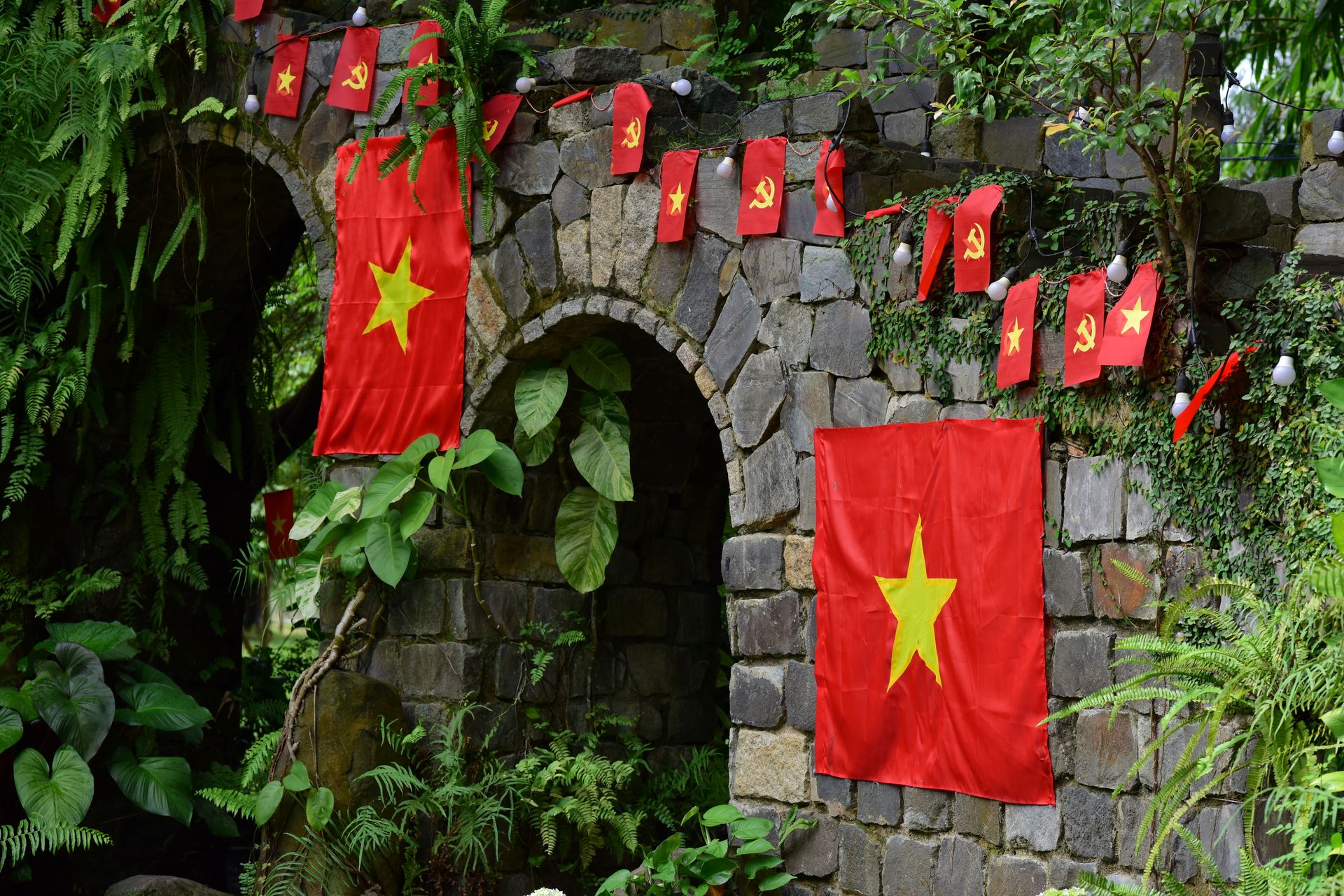 Vibrant Vietnamese flags decorate a stone arch surrounded by lush greenery in an outdoor setting.