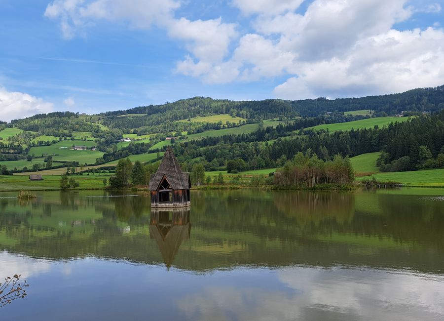 Scenic view of a wooden house on a lake with lush hills in Steiermark, Austria.