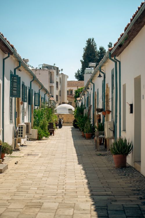 Free Quaint narrow street lined with white buildings and plants under a clear blue sky. Stock Photo