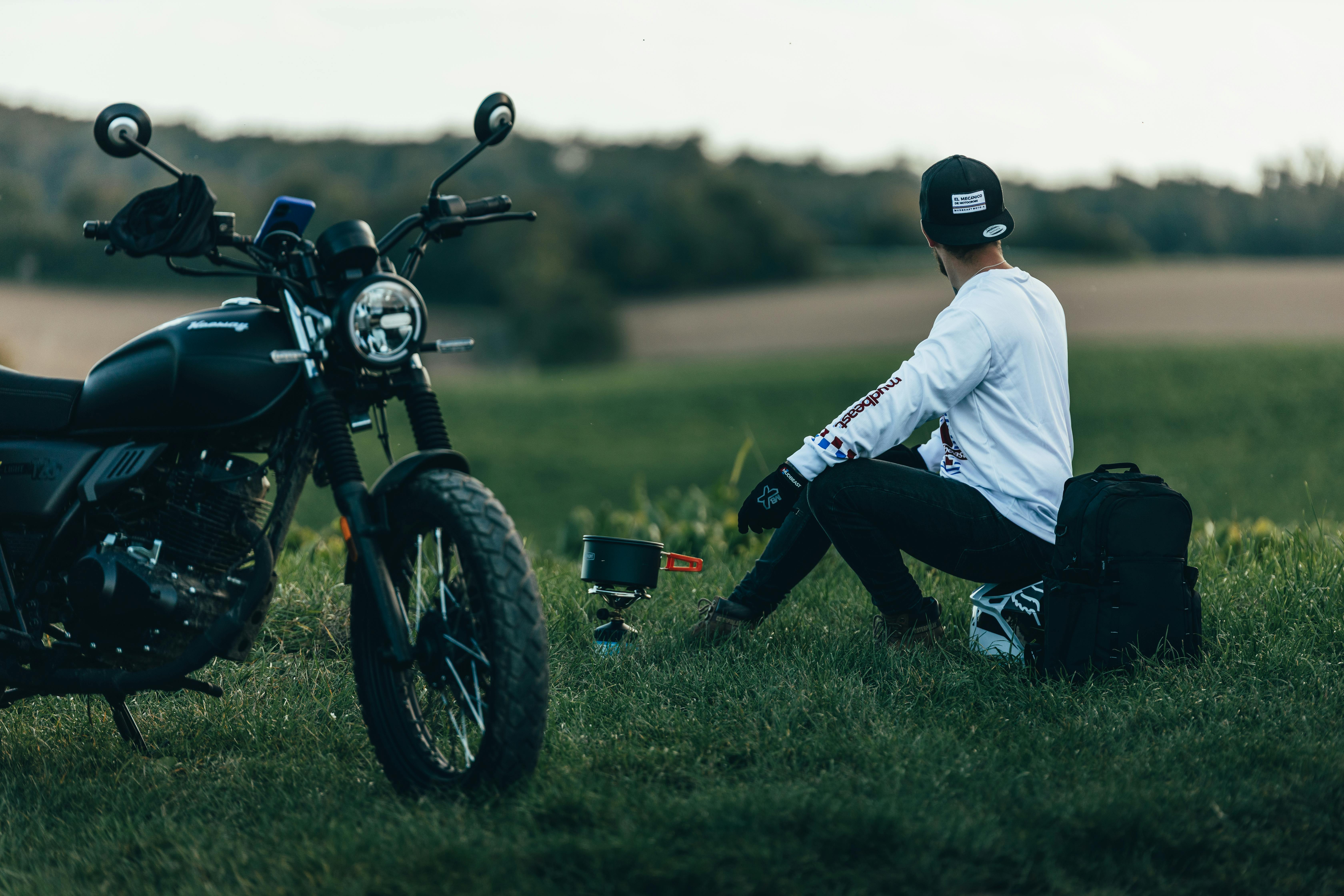 A man in casual attire sits by a motorcycle in a scenic, open field, enjoying a camping setup.