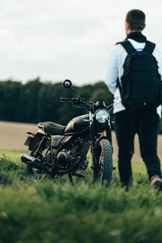 A man with a backpack stands beside a parked motorcycle in a rural setting, ready for adventure.