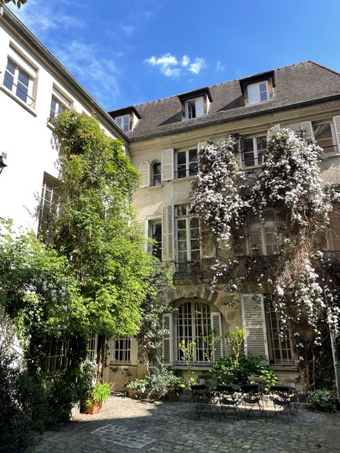 Lush greenery and flowers adorn a charming historic courtyard under a bright blue sky.