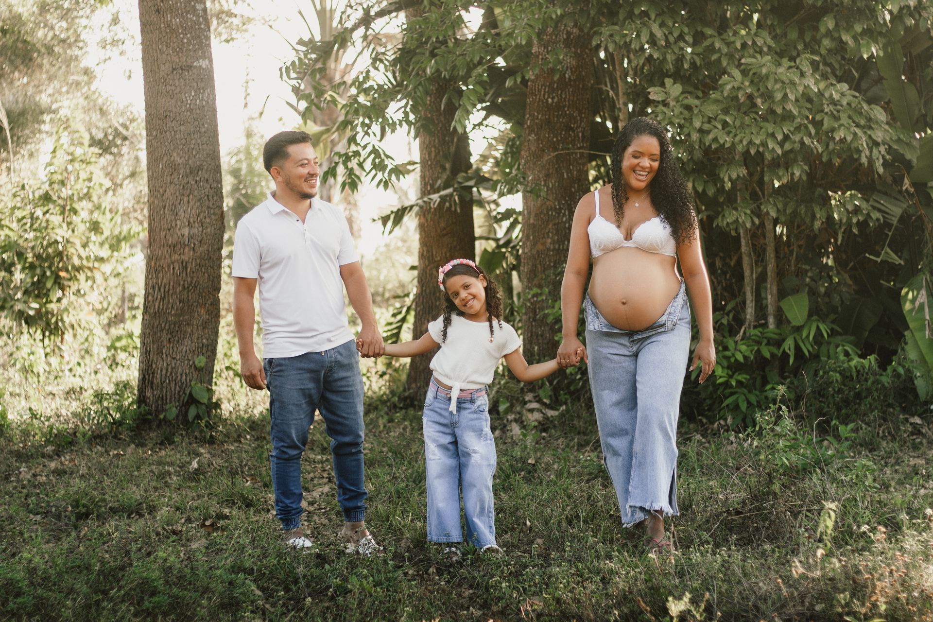 A joyous family portrait featuring a pregnant woman, man, and child holding hands in a lush setting.
