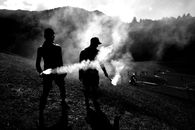 Silhouetted figures with smoke flares in a mountain landscape during daytime.