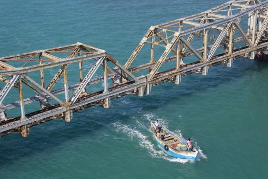 A fishing boat navigates beneath the historic rusty Pamban Bridge in clear blue waters.