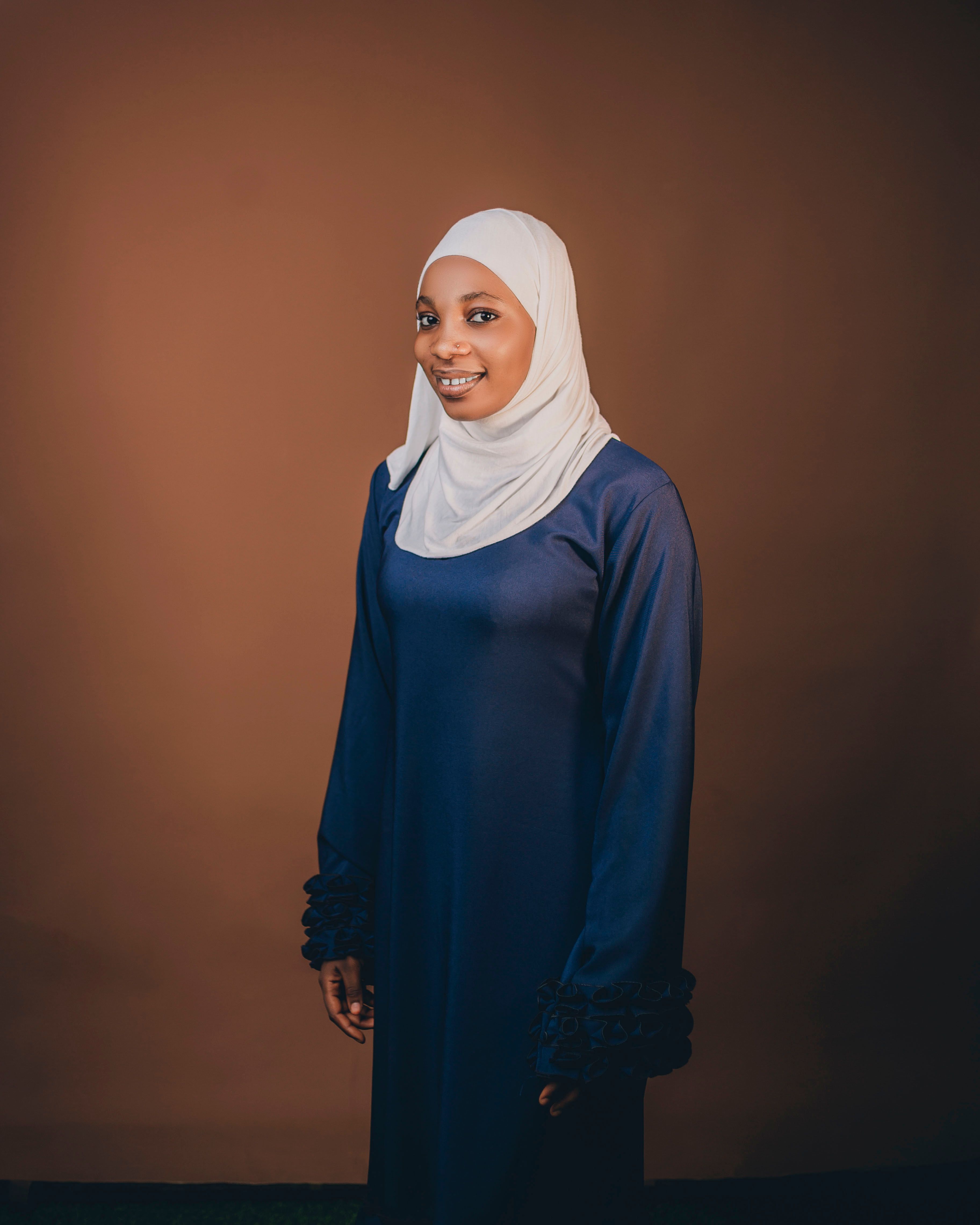 Elegant portrait of a woman wearing a hijab and blue dress against a brown background.