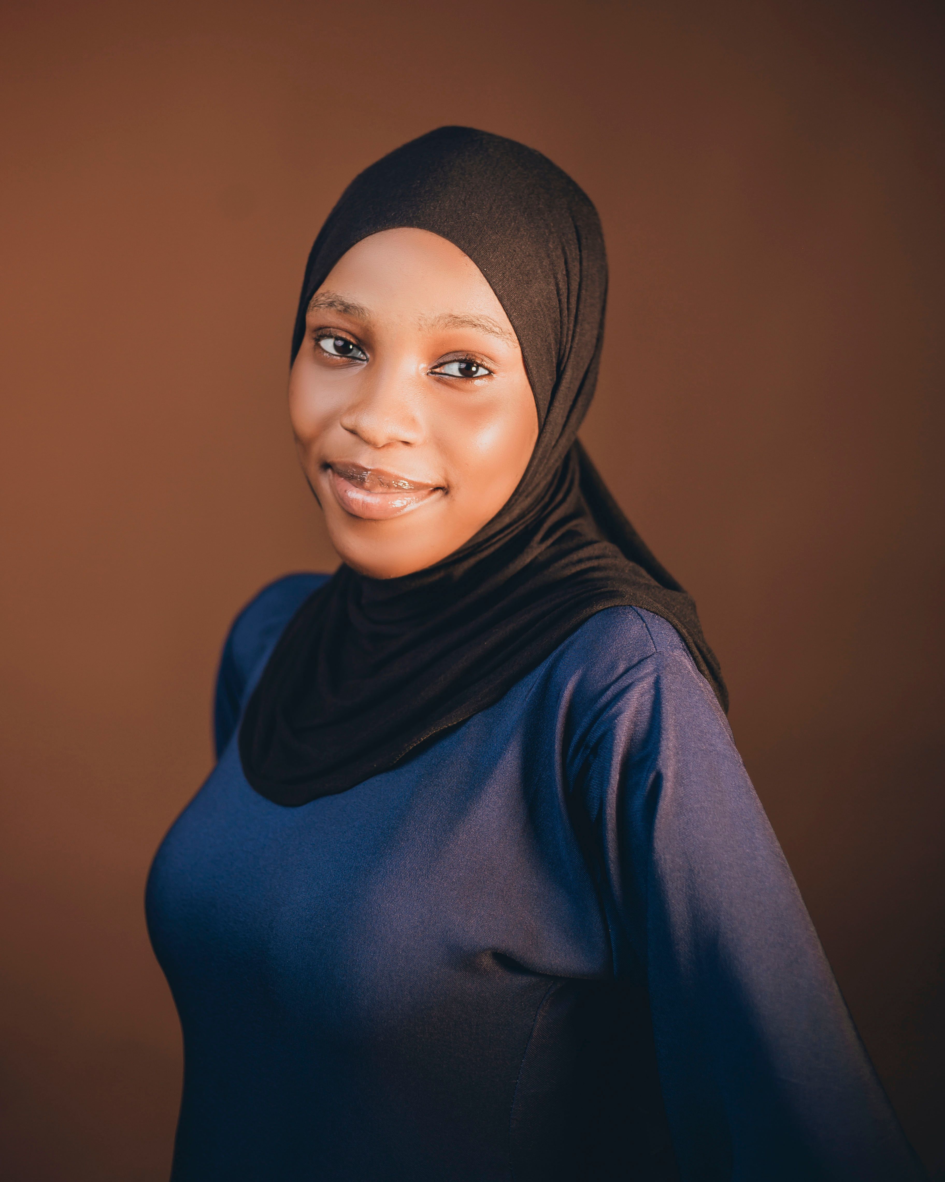 A confident woman in black hijab, smiling against a brown background.