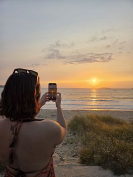 Woman photographs a vibrant beach sunset with her phone.