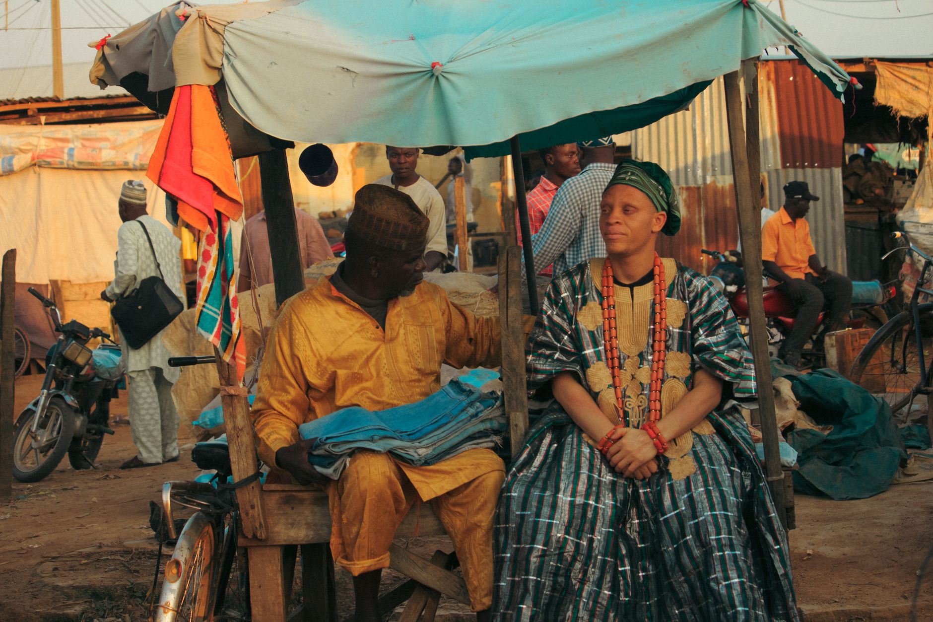 Two men in traditional attire at a bustling African street market. Vibrant colors and cultural garments.