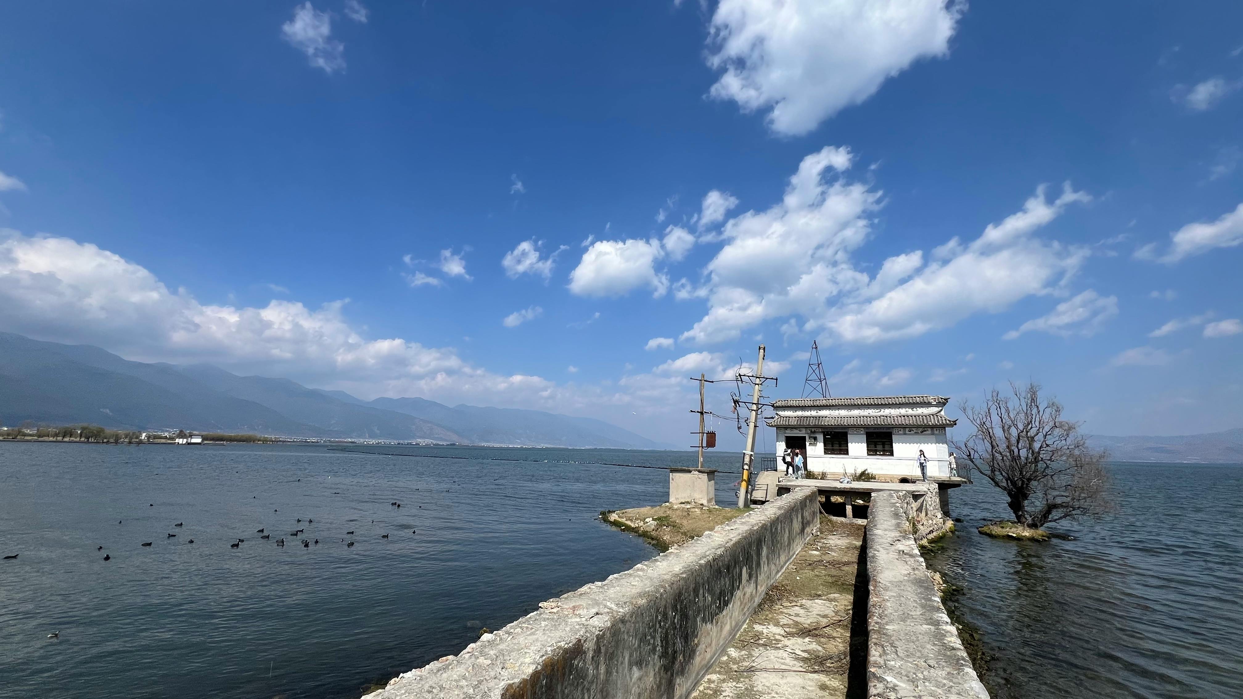 Stunning view of Erhai Lake with mountains, pier, and a house under a bright sky.