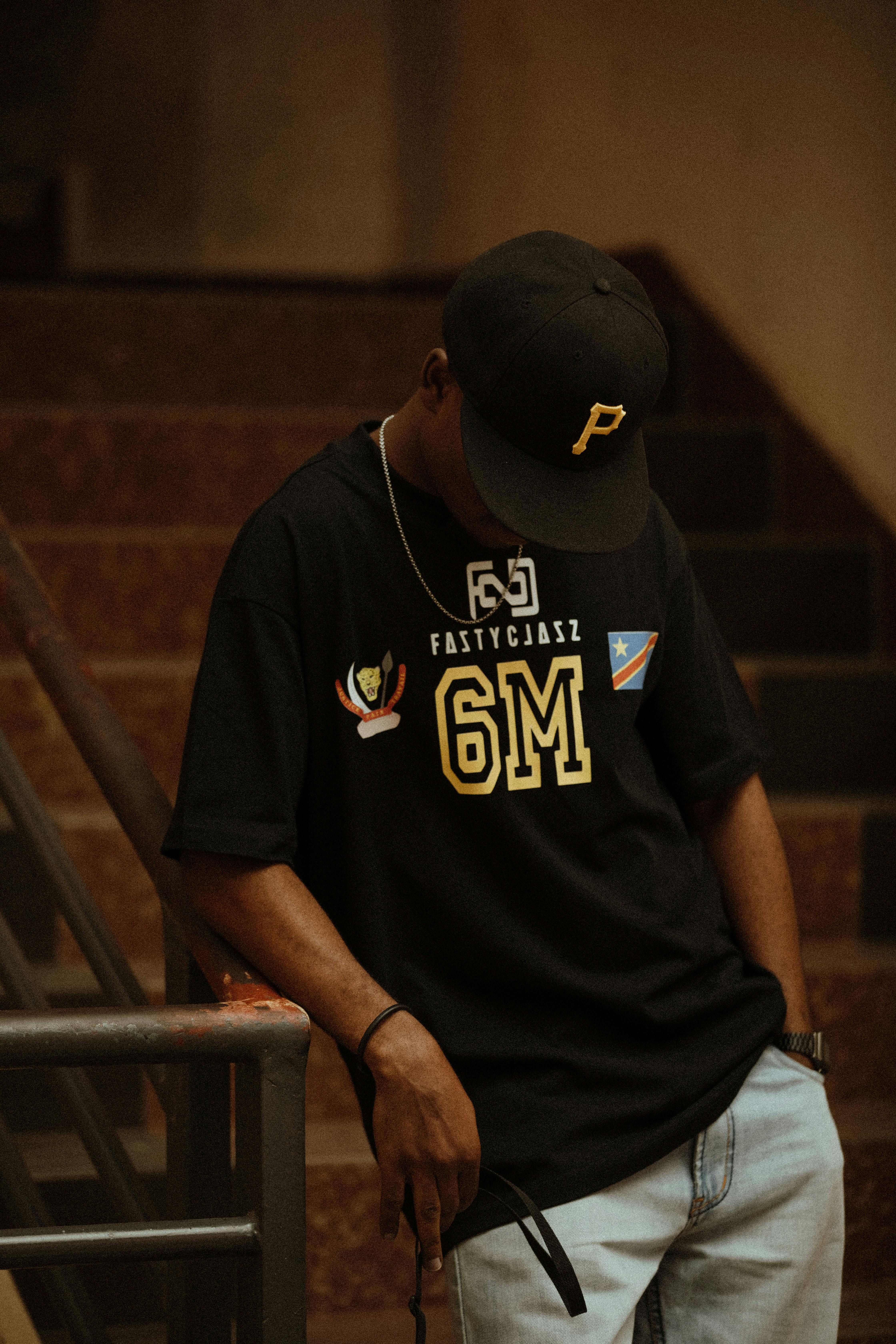 Young man in urban fashion leaning on staircase, wearing a graphic tee and baseball cap.
