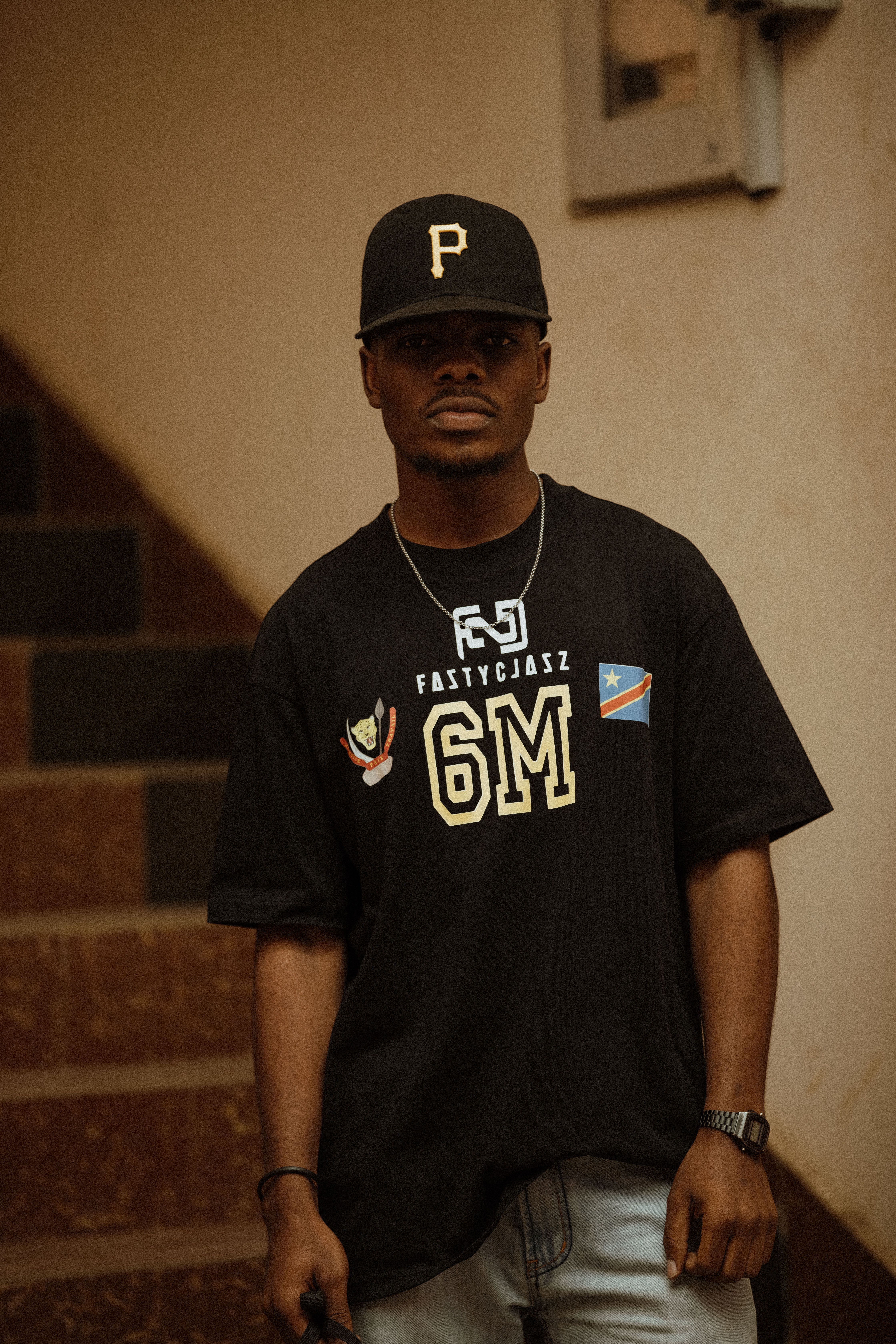 Portrait of a man wearing a black cap and graphic T-shirt, standing by stairwell indoors.