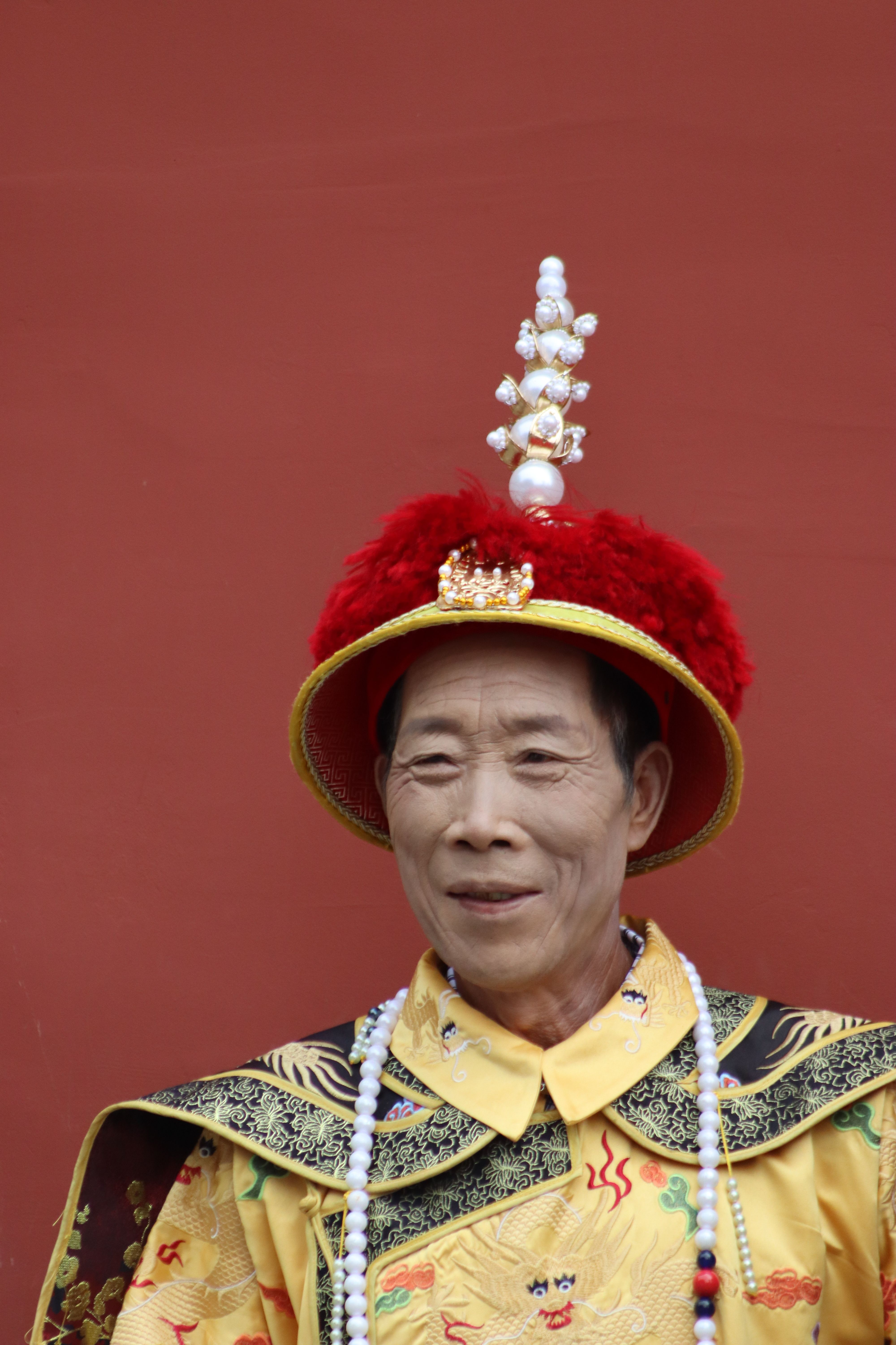 Portrait of an adult man in traditional Chinese attire with ornate headpiece against a red background.