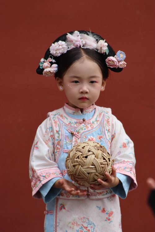 Free Young girl in traditional Chinese clothing holding a wicker ball against a red background. Stock Photo