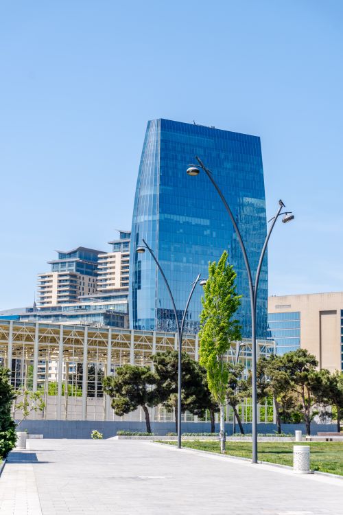Free A modern glass skyscraper in Baku, Azerbaijan, surrounded by green trees and clear blue sky. Stock Photo