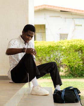 A young man sitting outdoors on a step, listening to music with earphones and a backpack beside him.