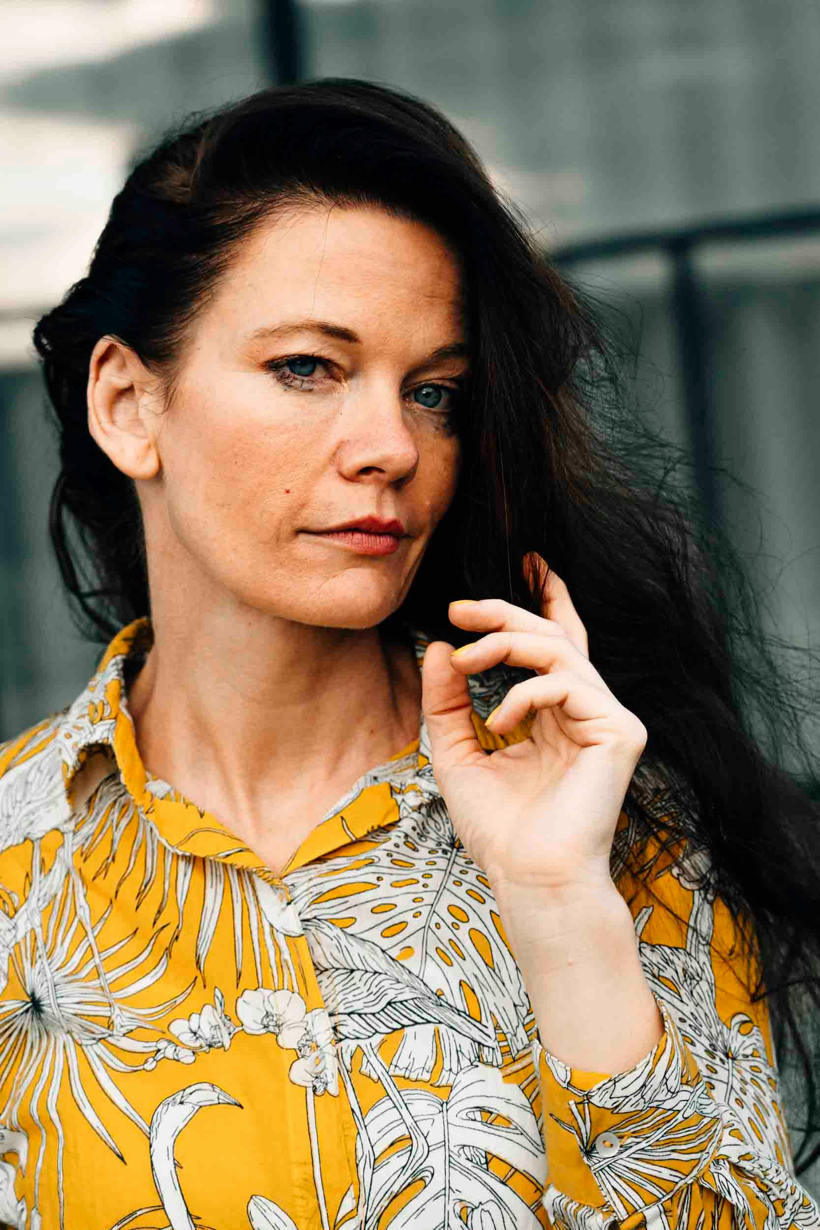Close-up portrait of a woman in a yellow shirt, captured outdoors with a confident expression.