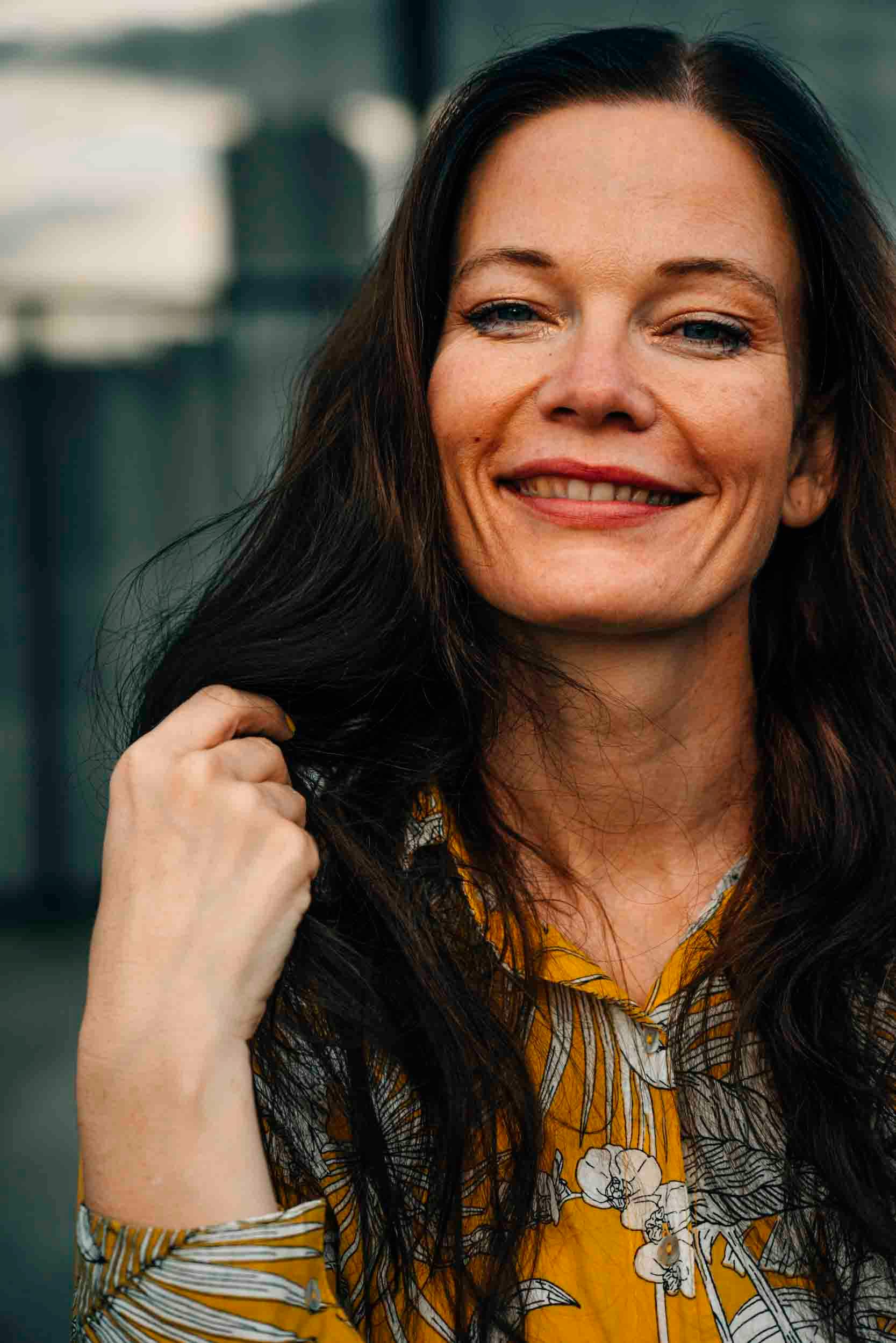 Charming woman smiling warmly in an outdoor setting, wearing a yellow shirt.