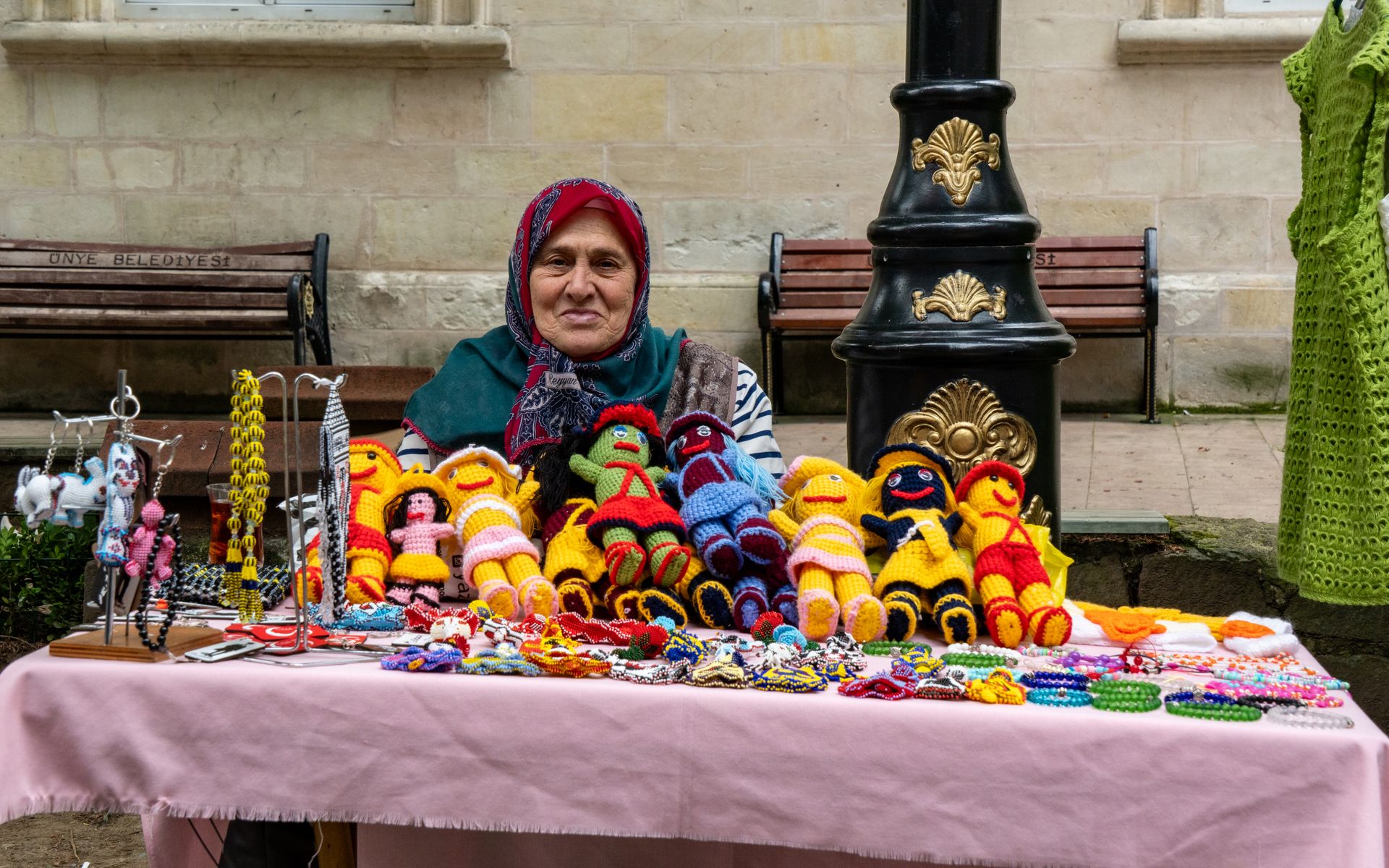 An elderly woman displays colorful handmade crafts at an outdoor market stall.