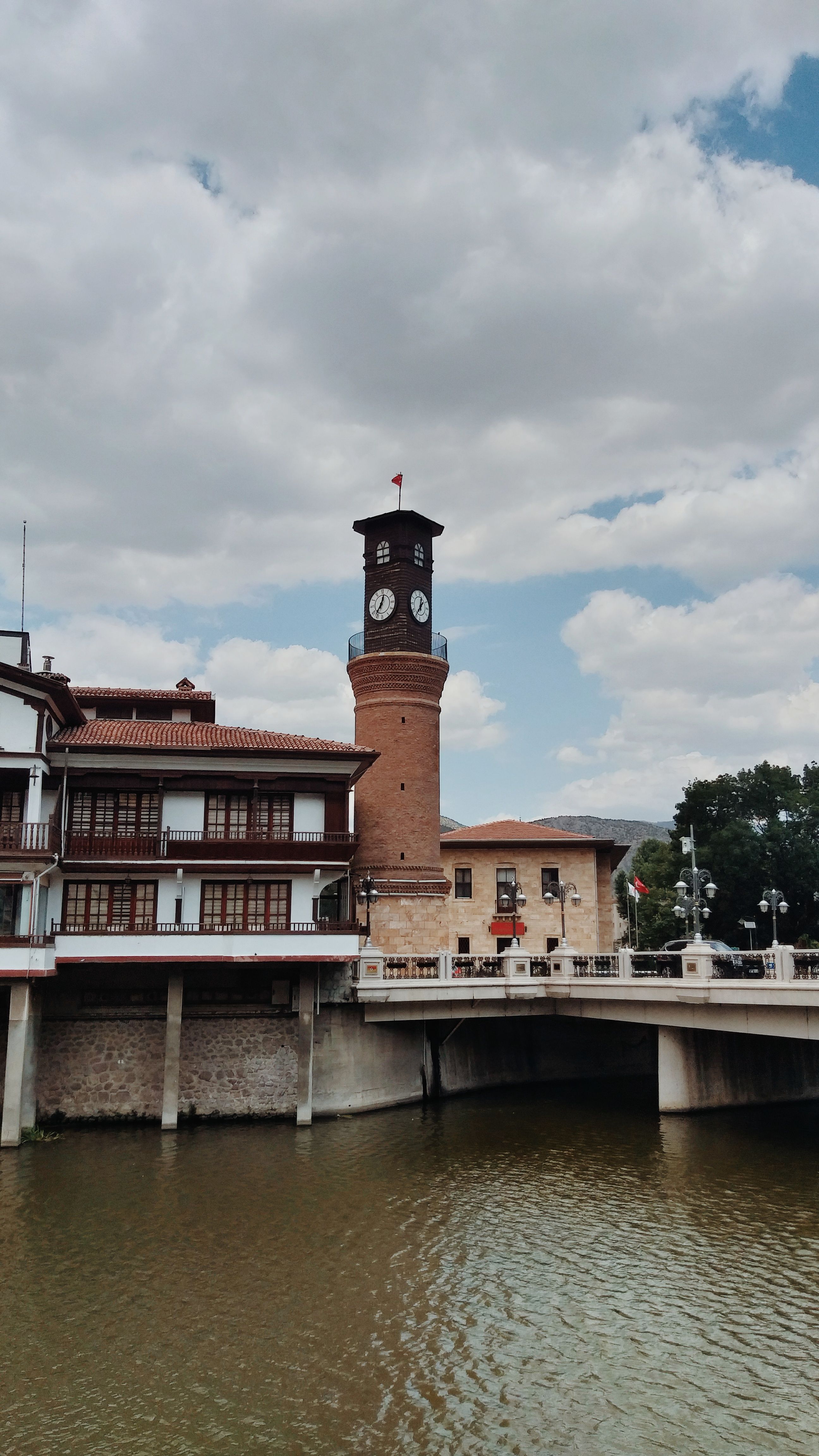 A scenic view of the historic clock tower and riverside buildings in Amasya, Türkiye, under a cloudy sky.