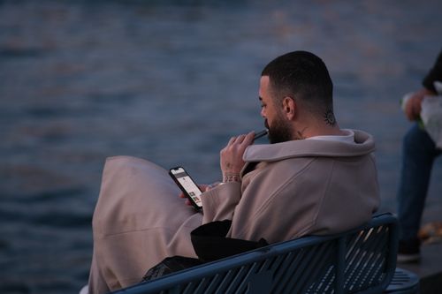 Free Person sitting by the water checking their phone and vaping, enjoying a tranquil moment. Stock Photo