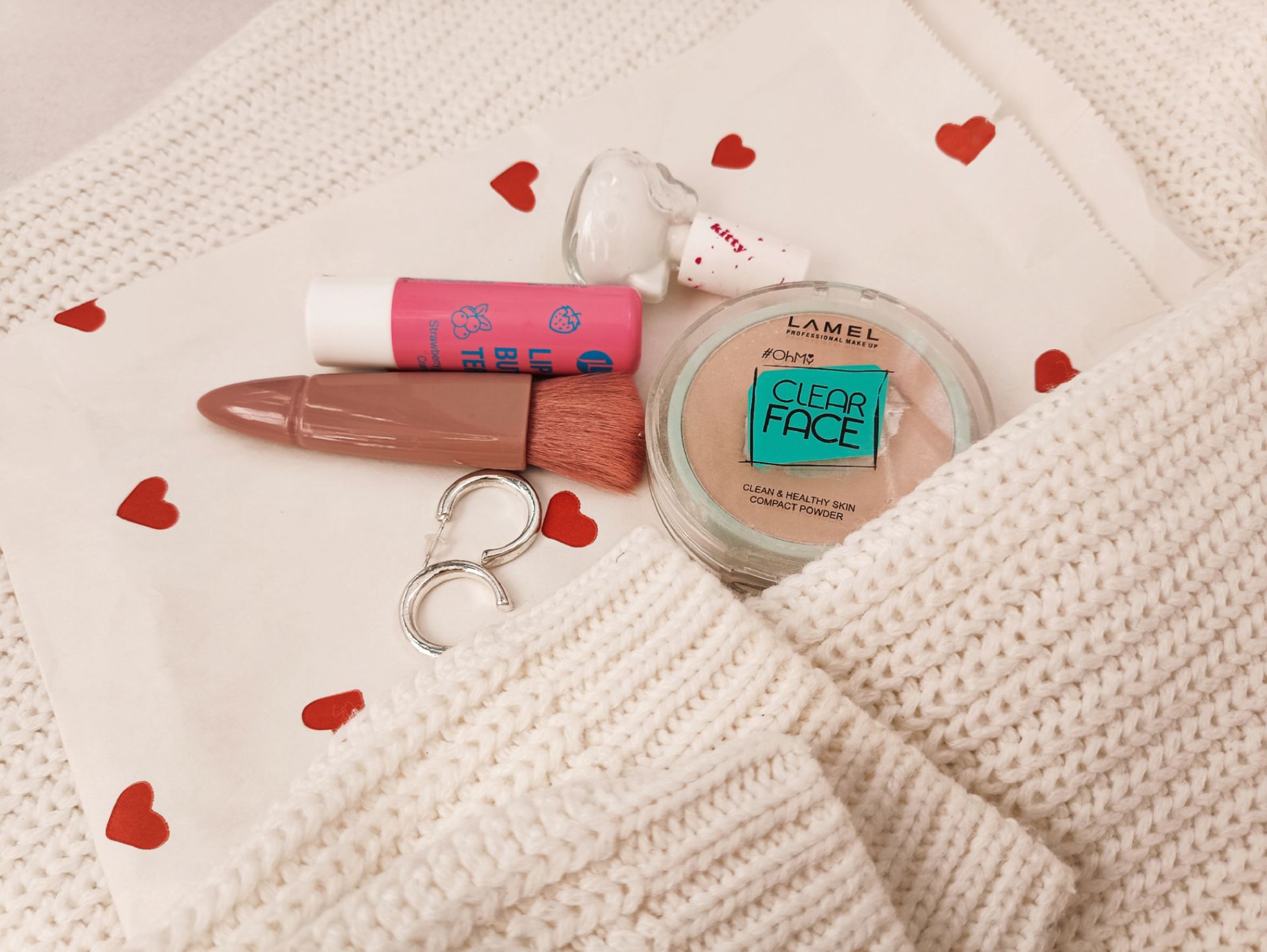 Stylish flat lay featuring makeup items, white knit, and heart-patterned cloth for beauty inspiration.