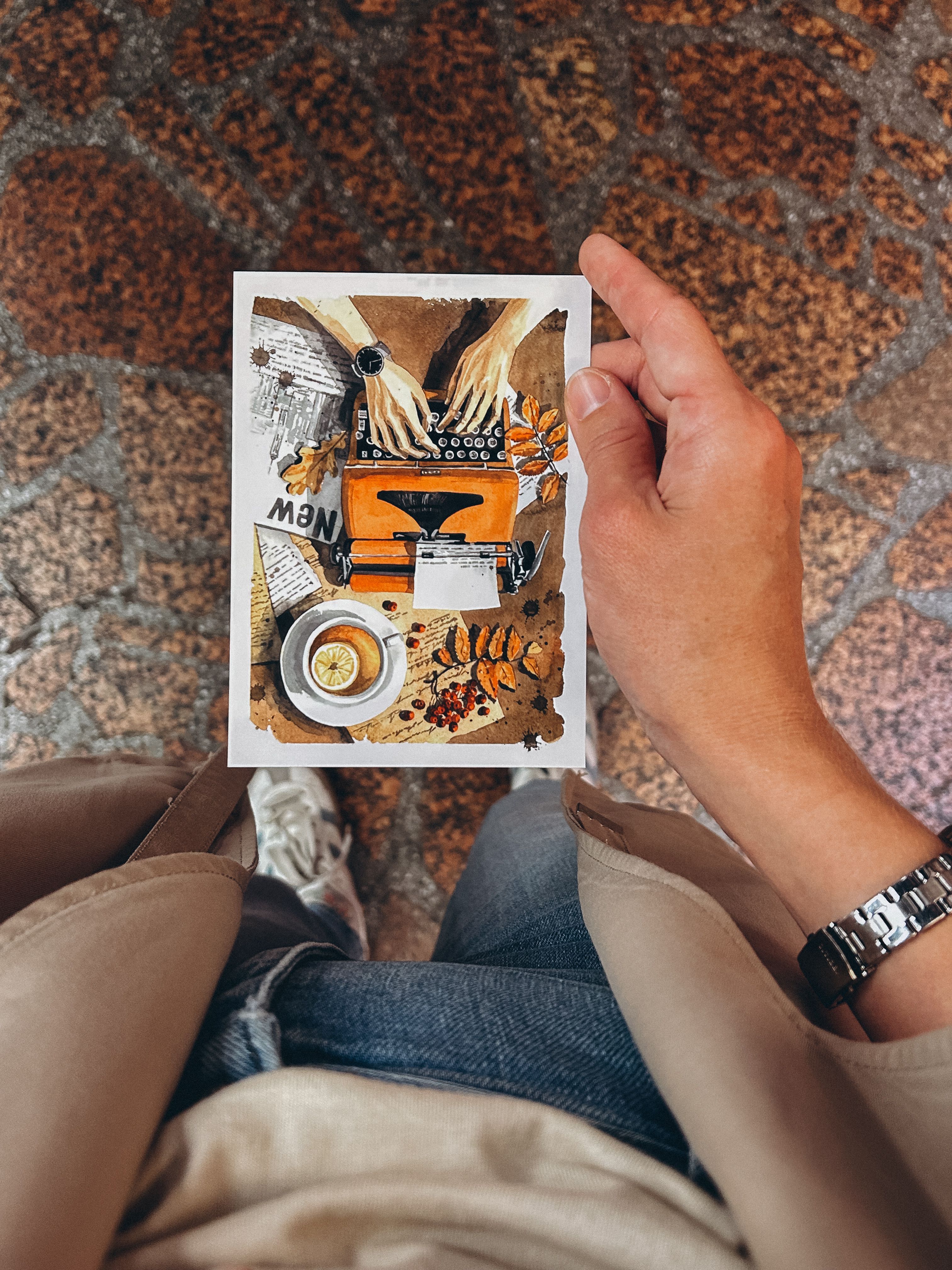 Hand holding a postcard with a typewriter and coffee illustration on a cobblestone street.