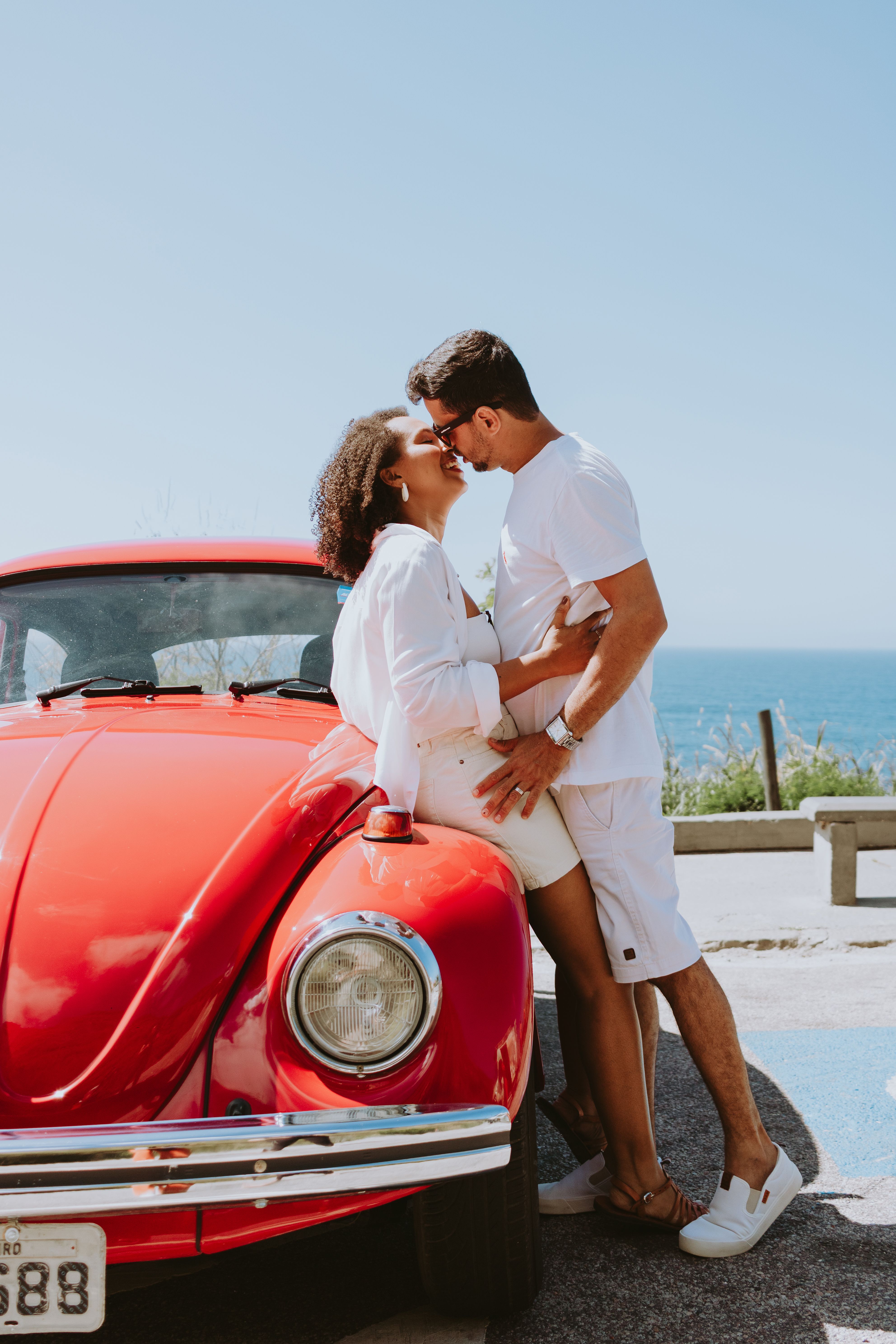 A couple embracing beside a classic red car with ocean views in Rio de Janeiro.