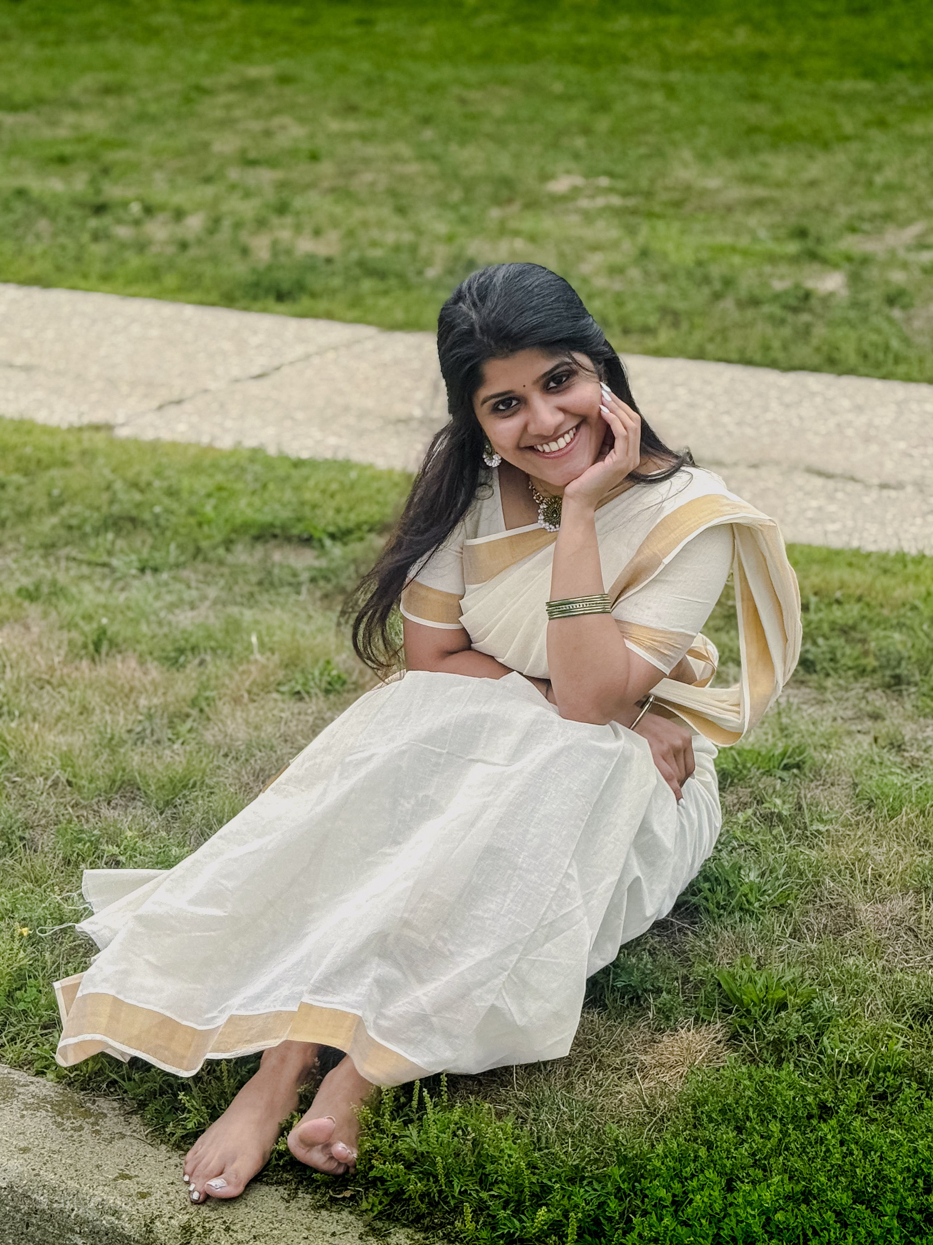 Woman wearing a white saree sits comfortably on the grass, smiling outdoors.