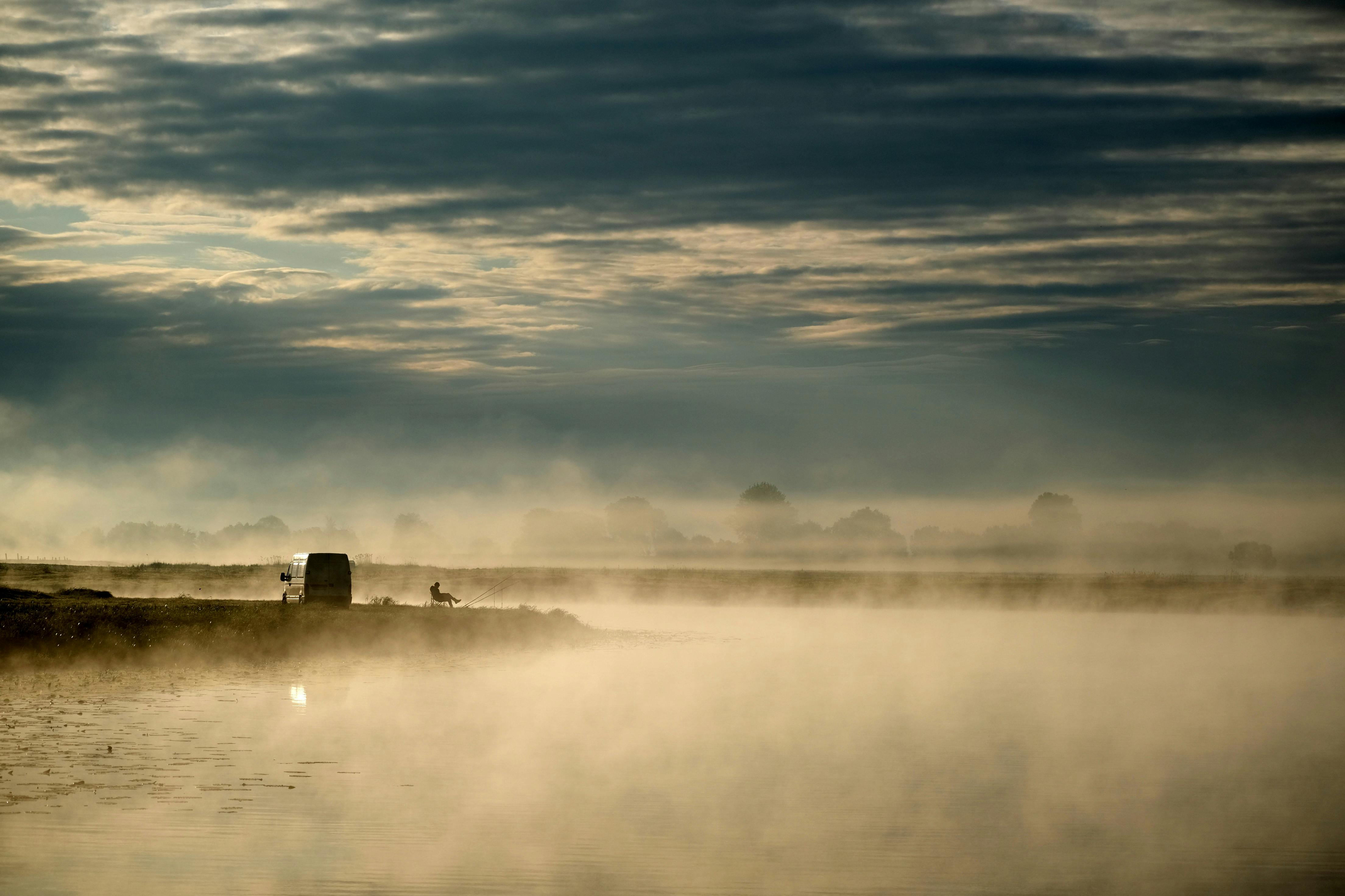 A solitary fisherman amidst morning mist on a tranquil lake enhances the serene atmosphere.