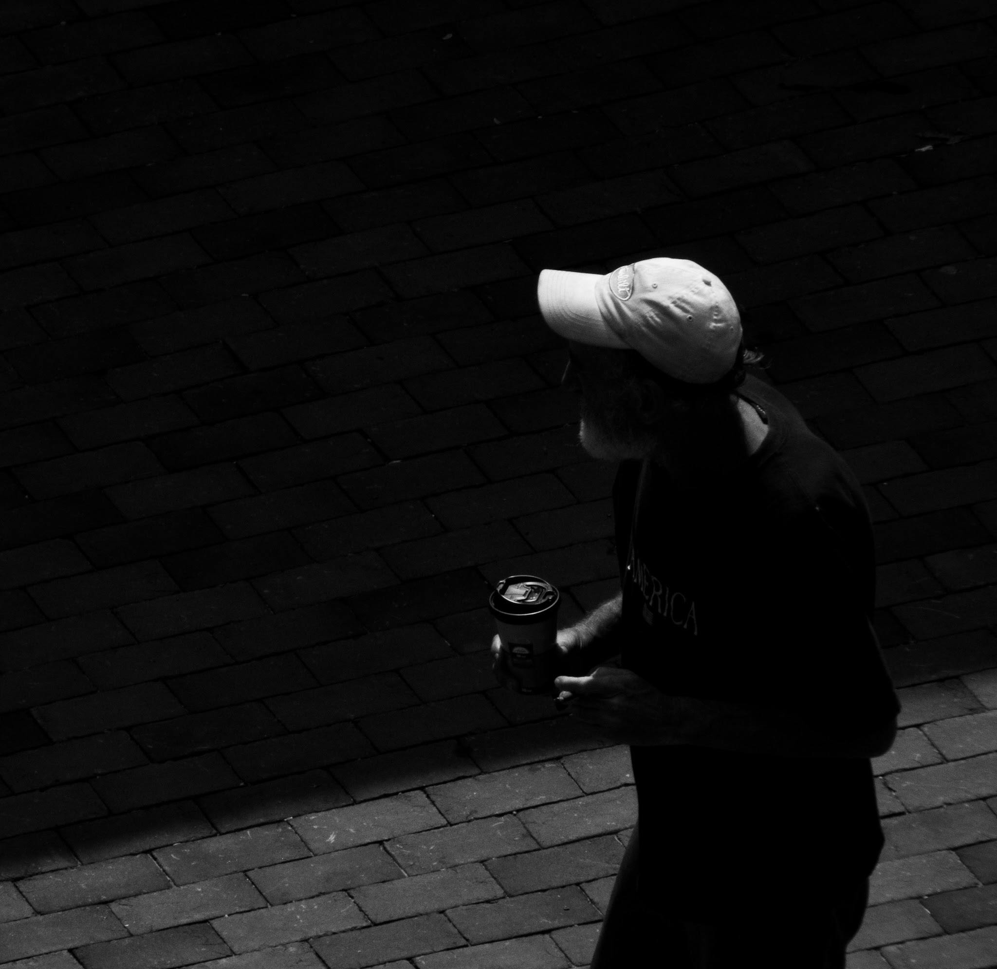 A dramatic black and white photo of a man walking on a sidewalk holding a coffee cup.
