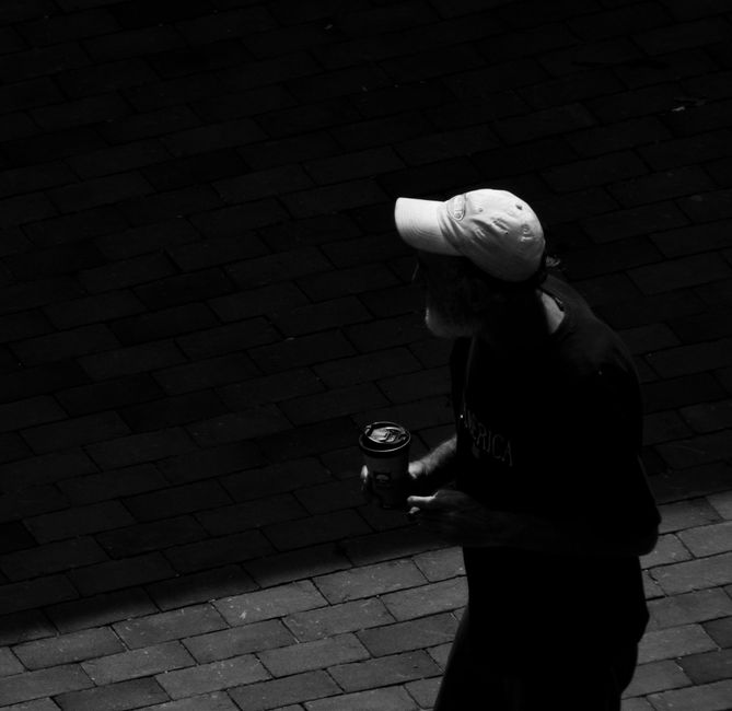 A dramatic black and white photo of a man walking on a sidewalk holding a coffee cup.