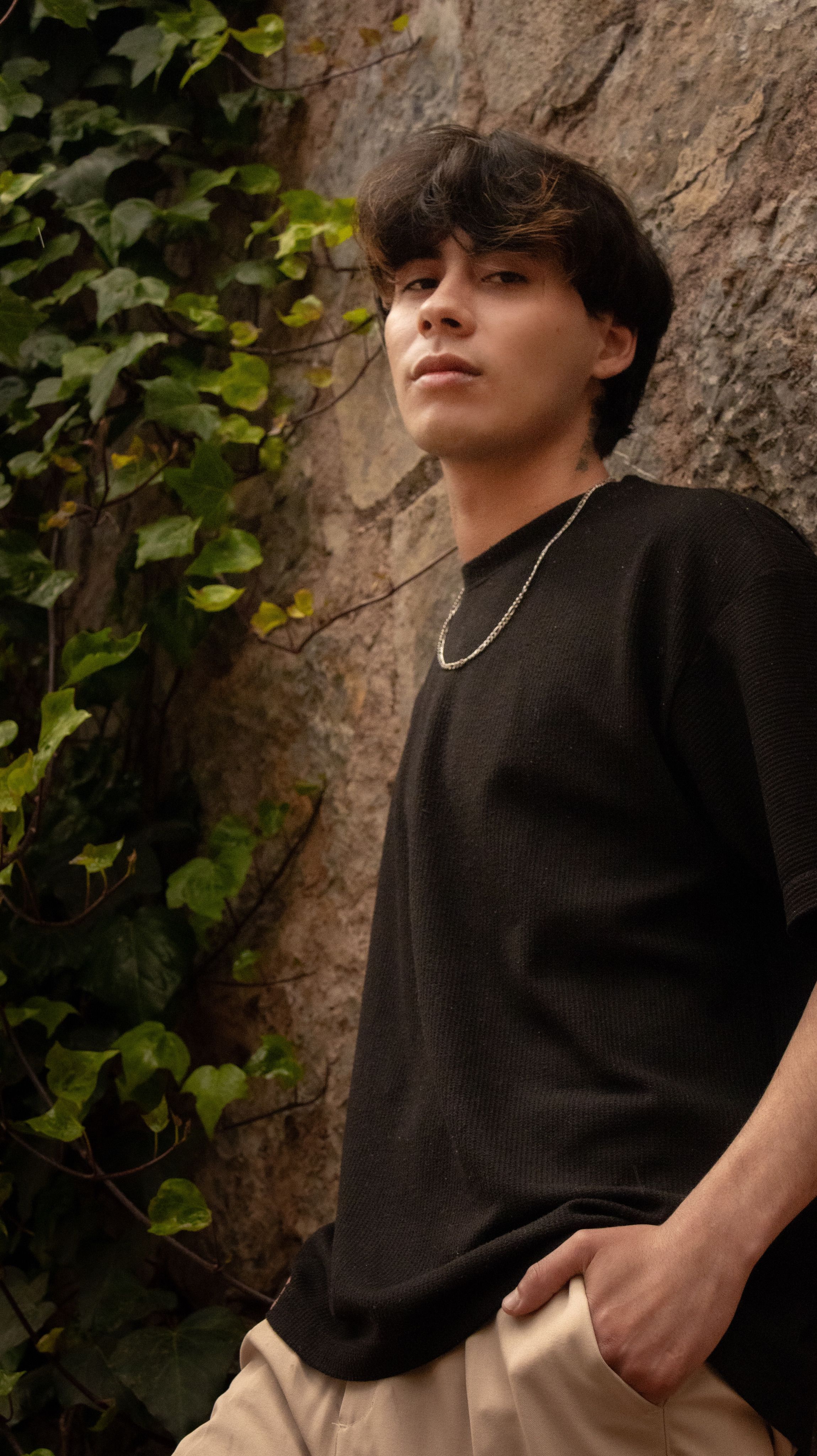 Stylish young man posing outdoors against a stone wall with greenery.