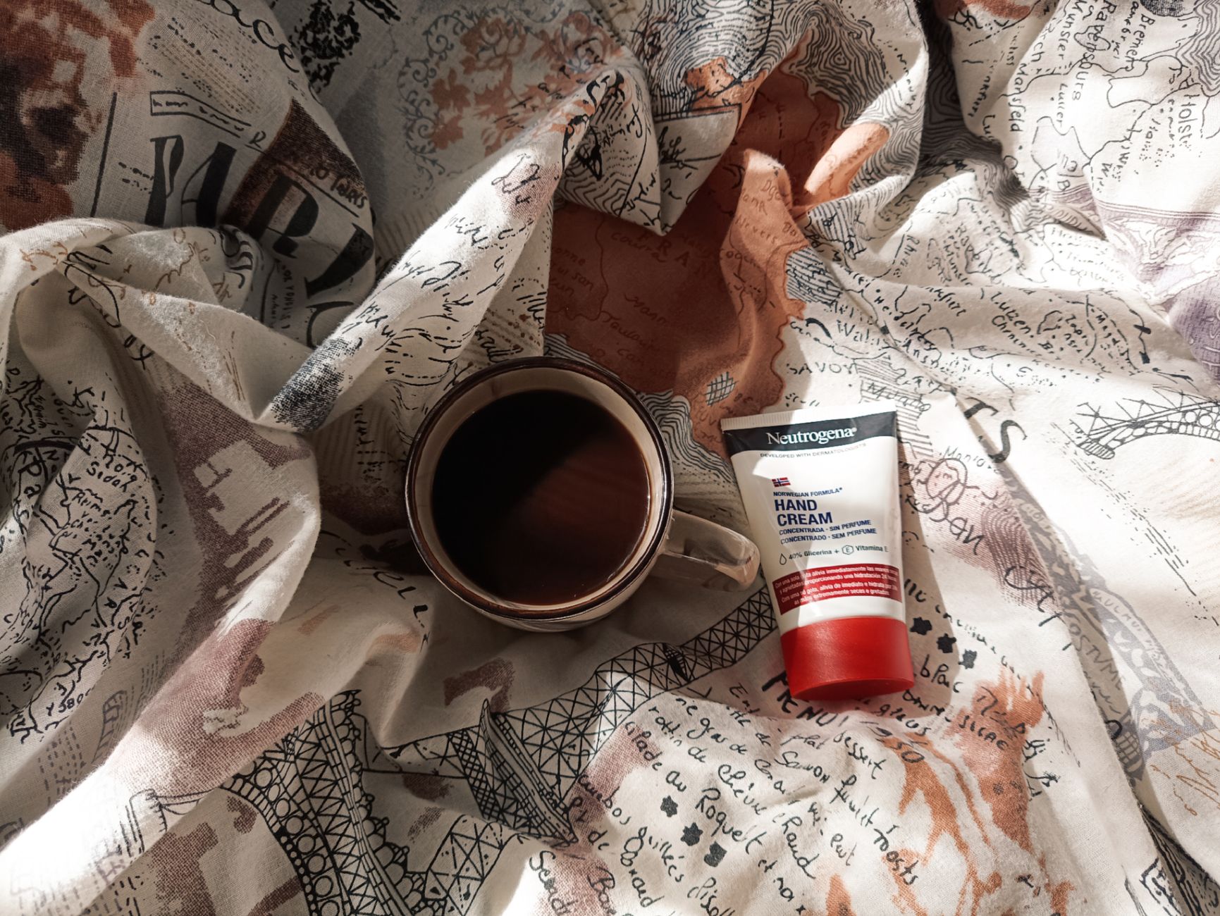 Flat lay of coffee and skincare on a patterned blanket, sunlit morning.