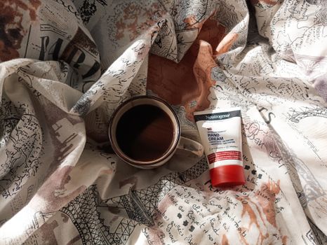 Flat lay of coffee and skincare on a patterned blanket, sunlit morning.
