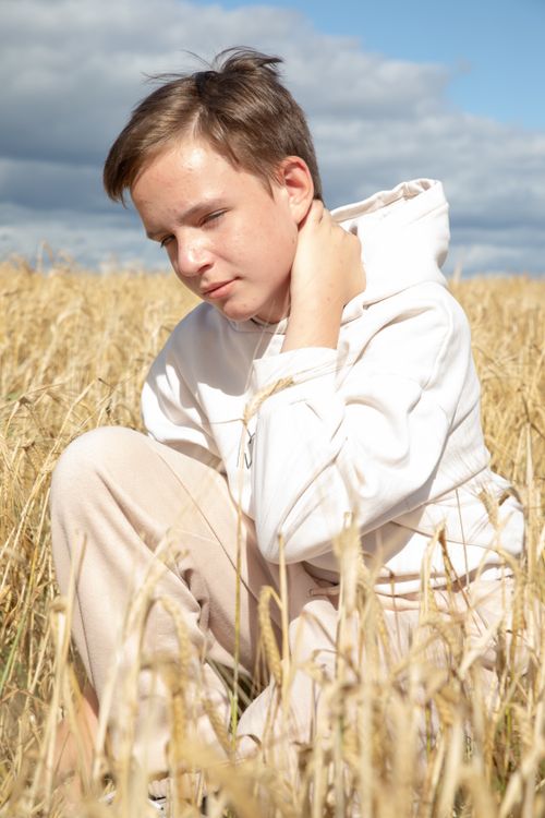 Free A teenager wearing a hoodie relaxes in a golden wheat field under a bright, cloudy sky. Stock Photo