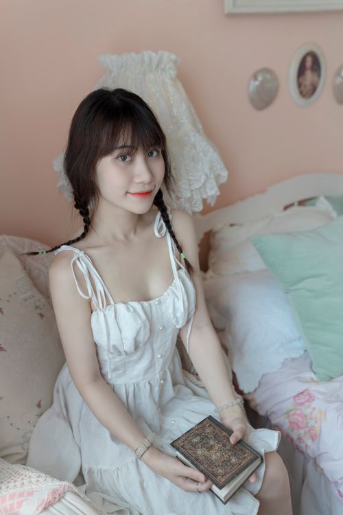 Free Young woman in a white dress sitting on a bed indoors, holding a book and smiling. Stock Photo