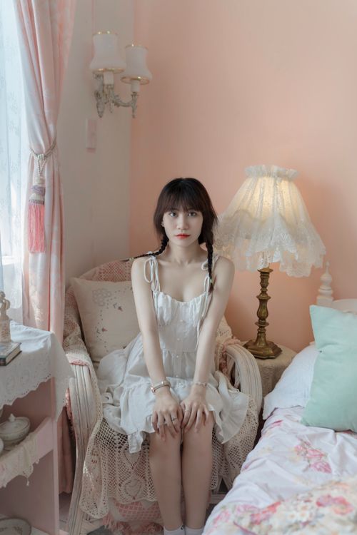 Free A young woman in a white dress sits in a pastel-colored bedroom, creating a soft and serene atmosphere. Stock Photo