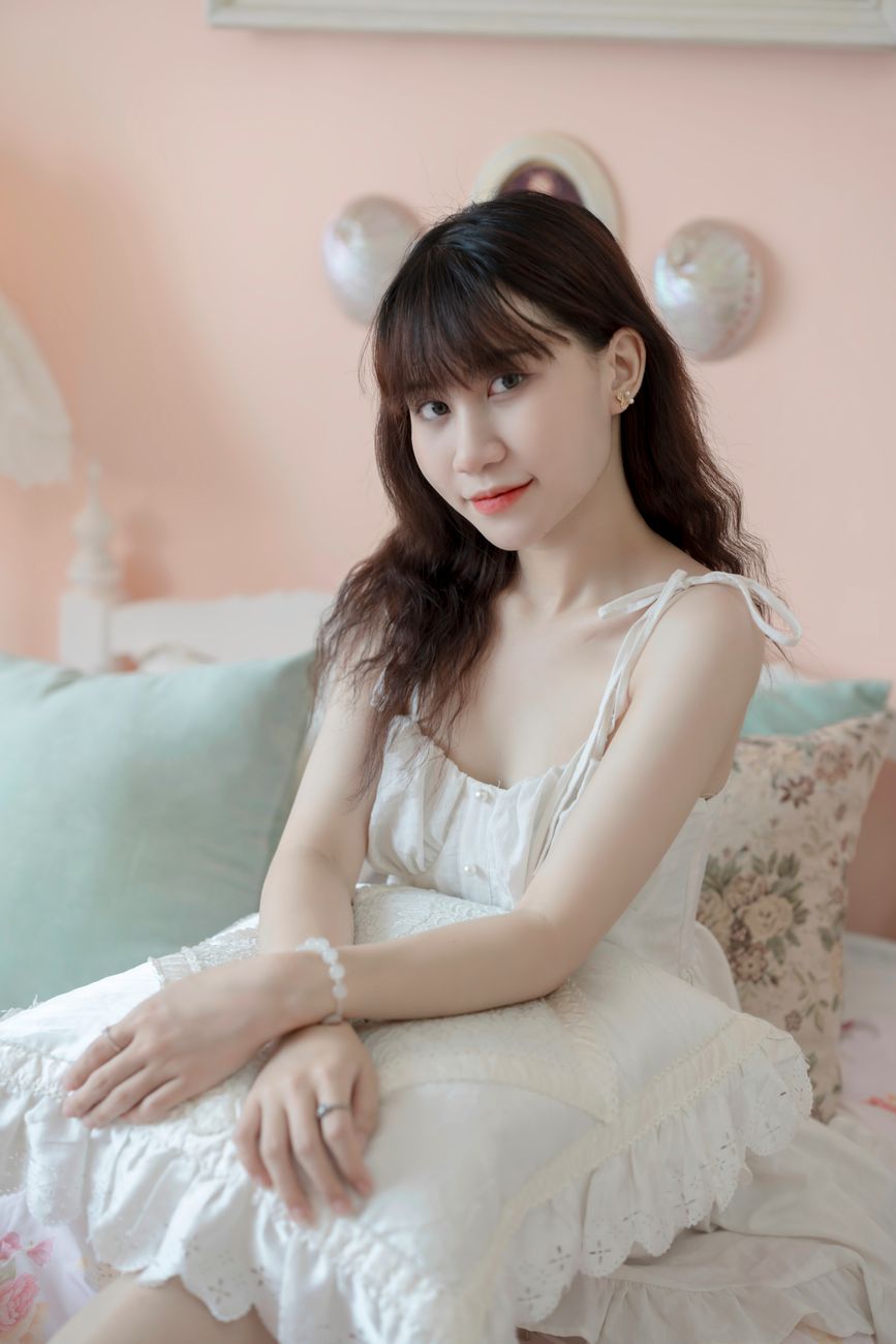 Charming young woman in a white dress sitting gracefully in a cozy, pastel-themed interior.