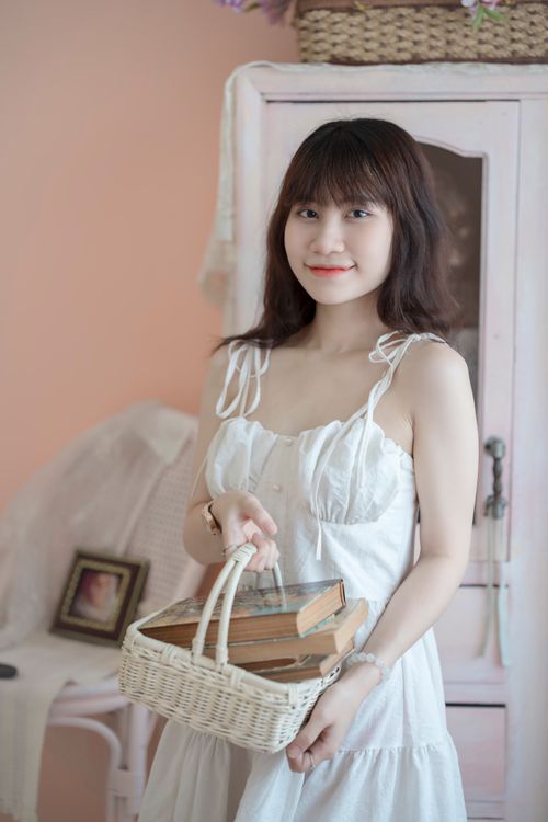 Free A young woman in a white dress holds a basket of books, standing in a cozy room with soft pastel decor. Stock Photo