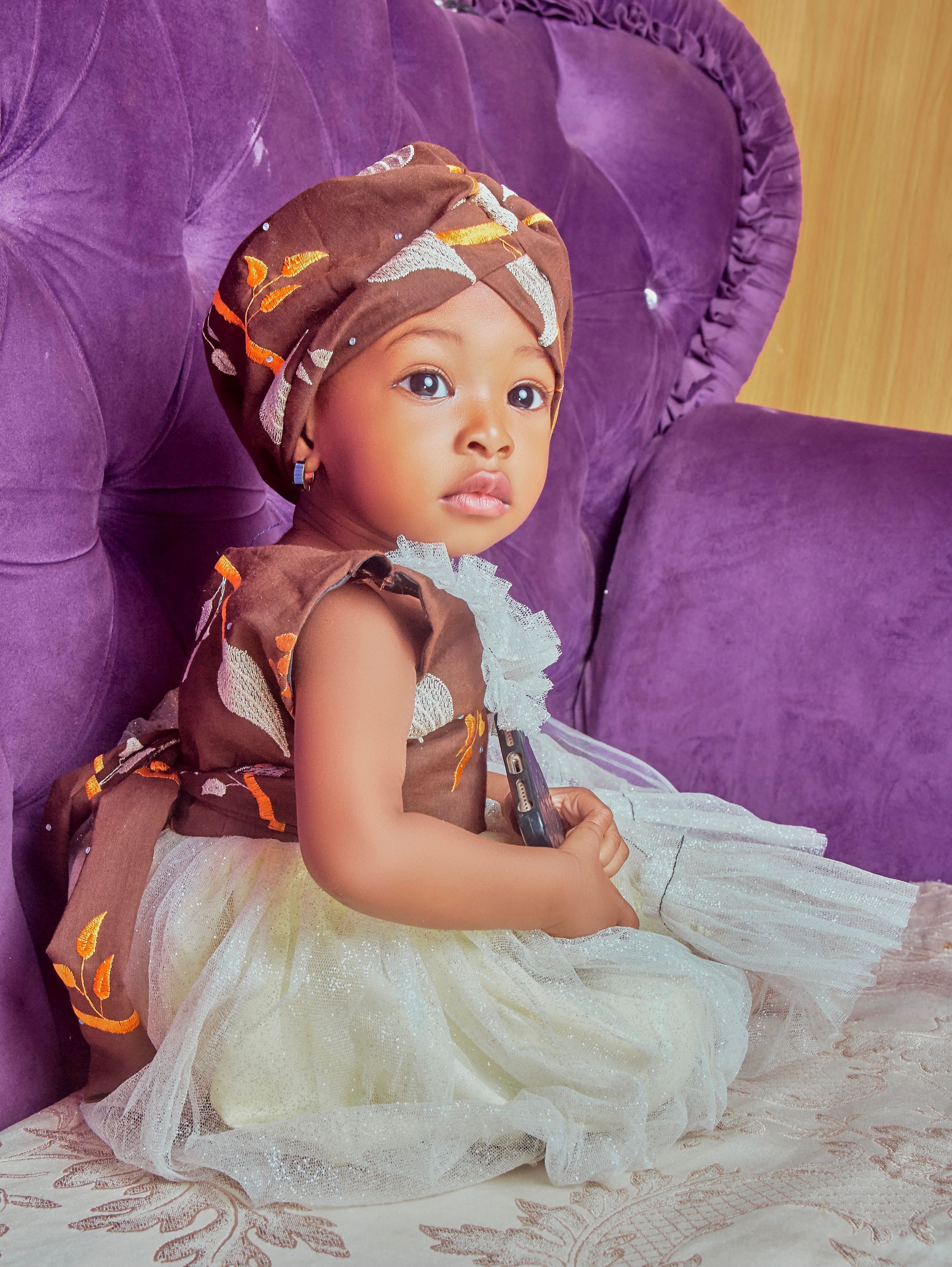 Charming baby in traditional African dress seated on a purple couch indoors.