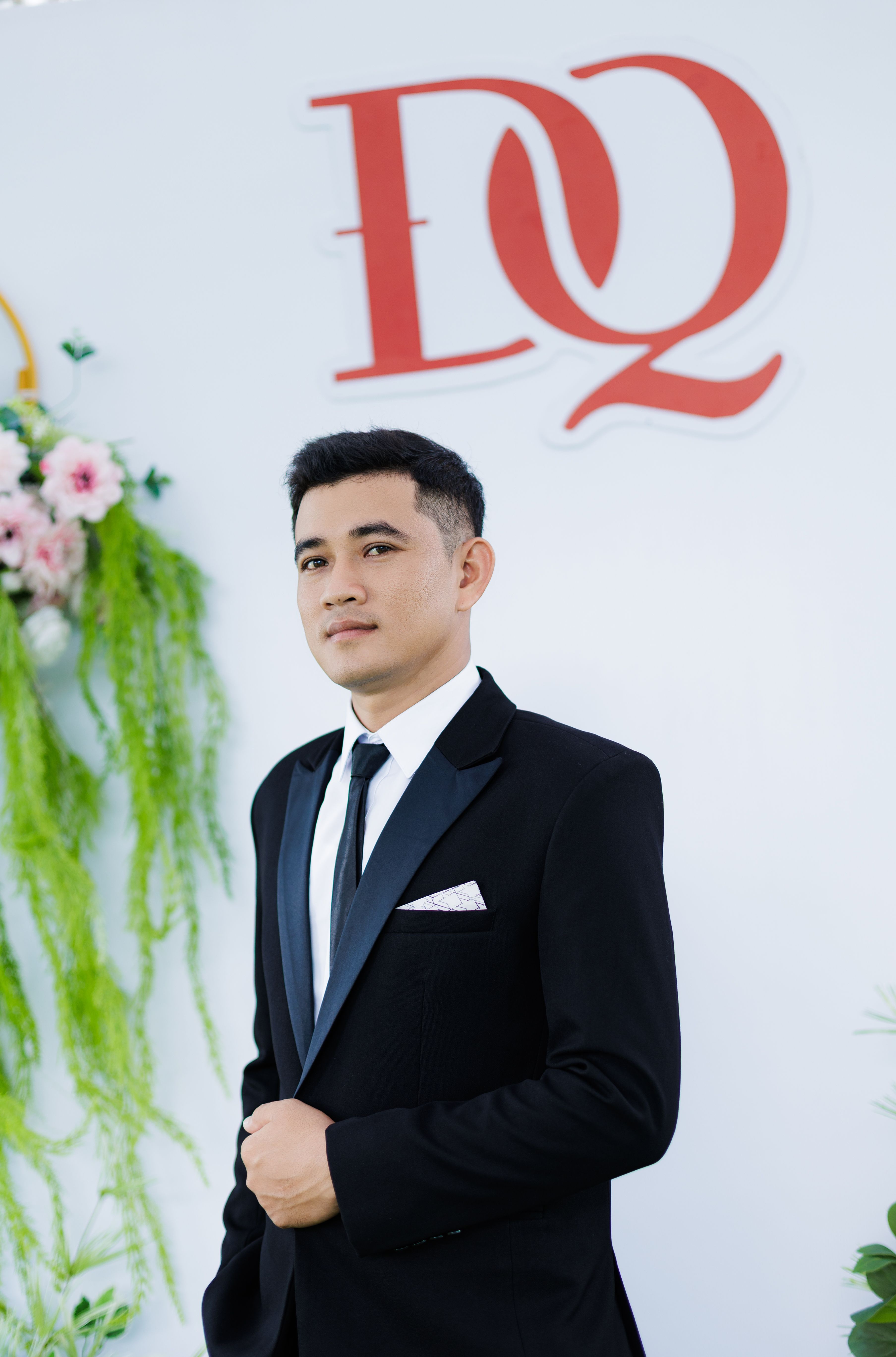 Handsome young man in a black suit stands confidently at a formal event with floral decor.