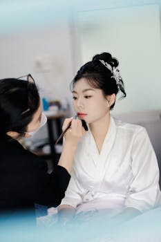 Asian bride in a white robe having her makeup done by a professional artist indoors.