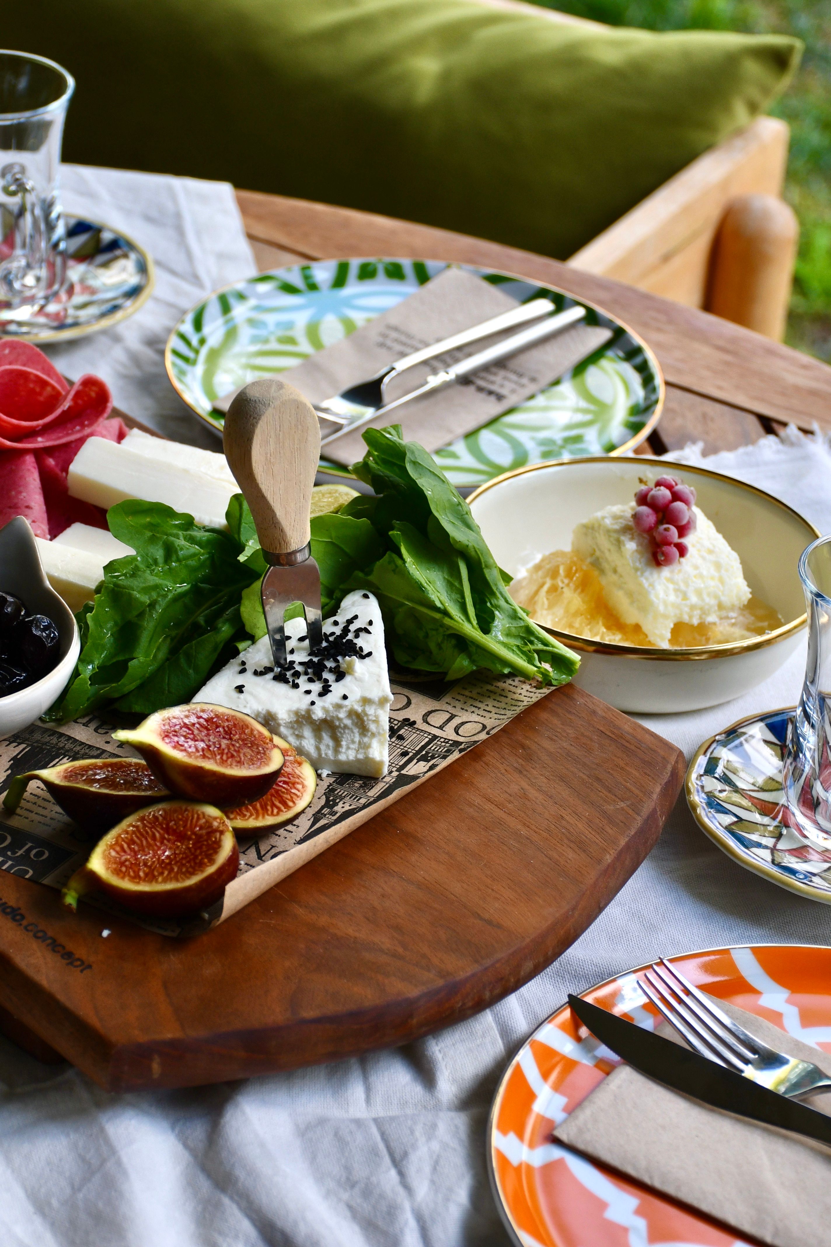 A stylish outdoor dining setup featuring cheese, figs, and leafy greens on a wooden board.