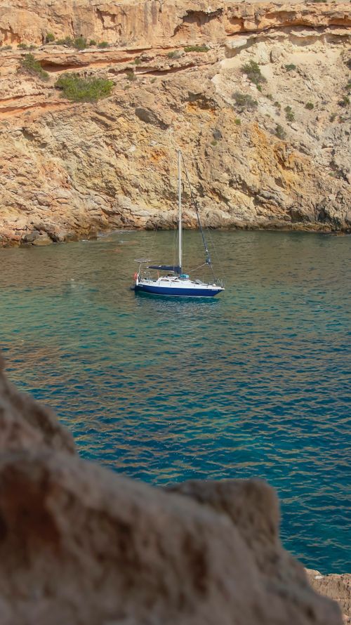 Free A serene sailboat anchored in a calm cove with rugged cliffs and clear blue water. Stock Photo