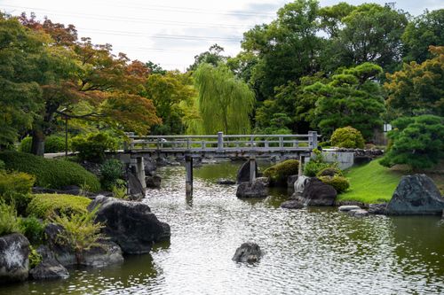 Free A serene view of a traditional Japanese garden with a rustic wooden bridge over a peaceful pond. Stock Photo